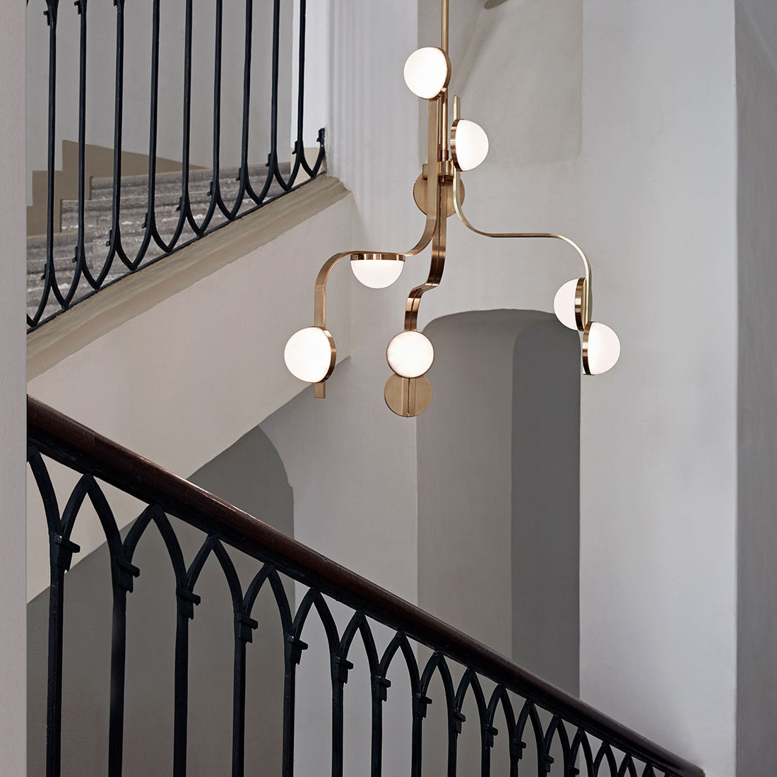 The Script Chandelier by Lobmeyr, featuring a gold frame and six frosted glass LED lights, graces the stairwell center. Black iron railings and white walls enhance this minimalist interior with a touch of Viennese Art Deco elegance.