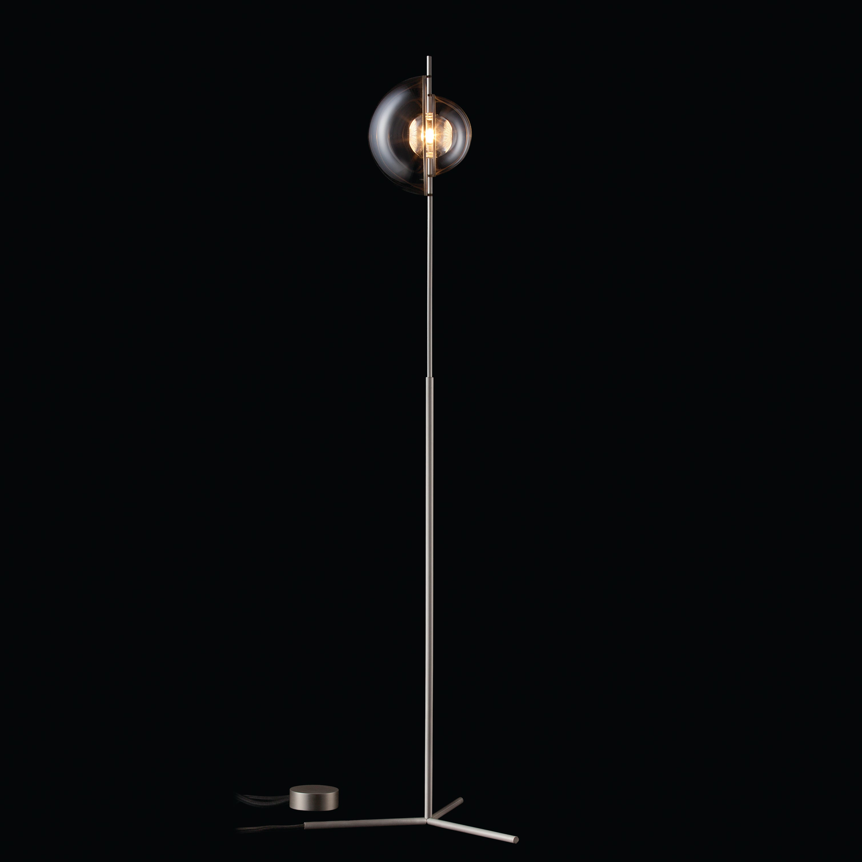 The Captured Floor Lamp by Lobmeyr is a contemporary piece with a sleek metallic stand and a glowing bulb encased in a glass sphere, all set against a black background.