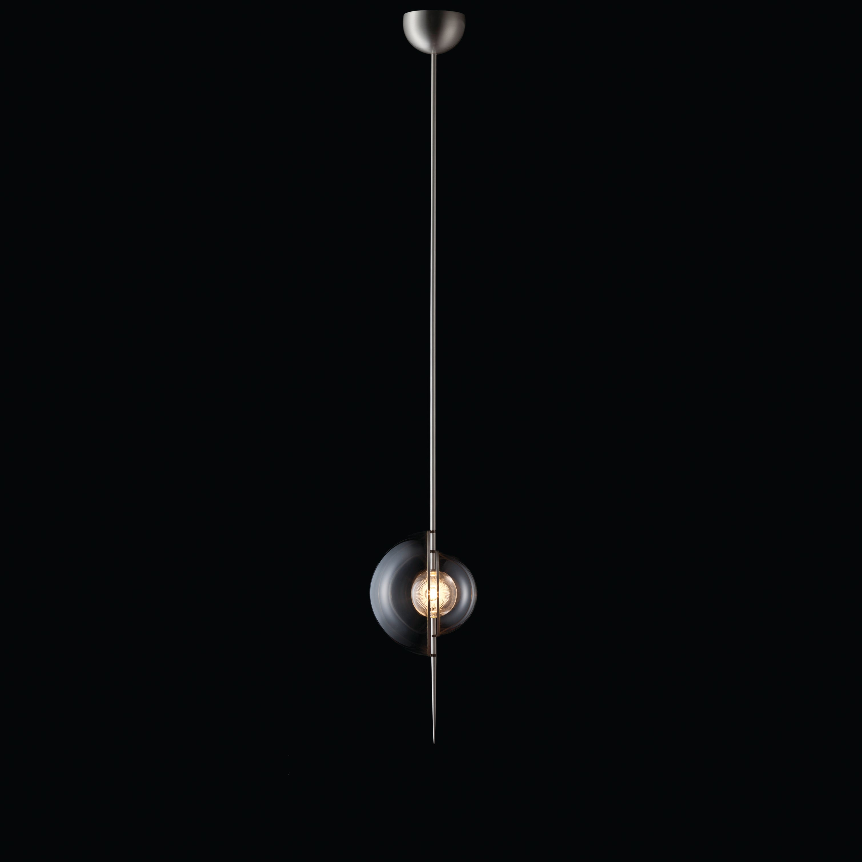 The Captured Pendant Lamp by Lobmeyr highlights minimalism with a long, thin metal rod and a lead-free crystal sphere encasing an exposed bulb set against a black background, exuding sleek elegance.