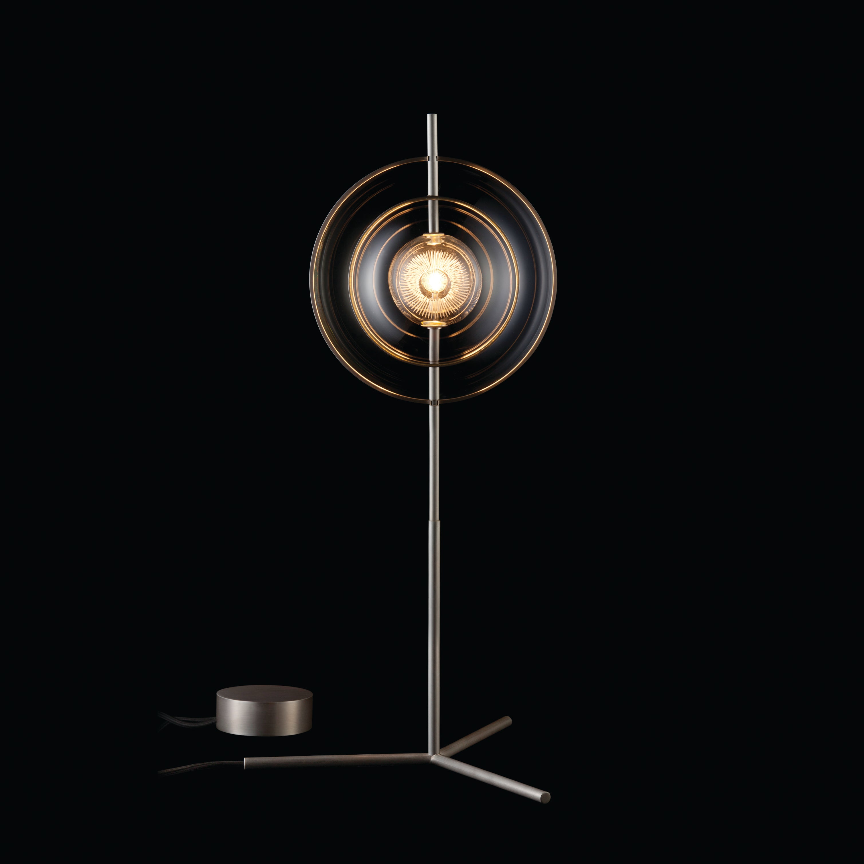 The Captured Table Lamp by Lobmeyr features a modern metallic design with a glass sphere encased in circular rings on a slender stand. Its elegant structure refracts light beautifully, creating an upscale ambiance against a dark background.