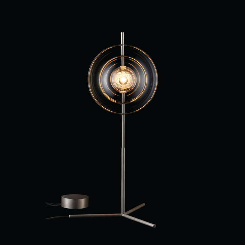 The Captured Table Lamp by Lobmeyr features a modern metallic design with a glass sphere encased in circular rings on a slender stand. Its elegant structure refracts light beautifully, creating an upscale ambiance against a dark background.