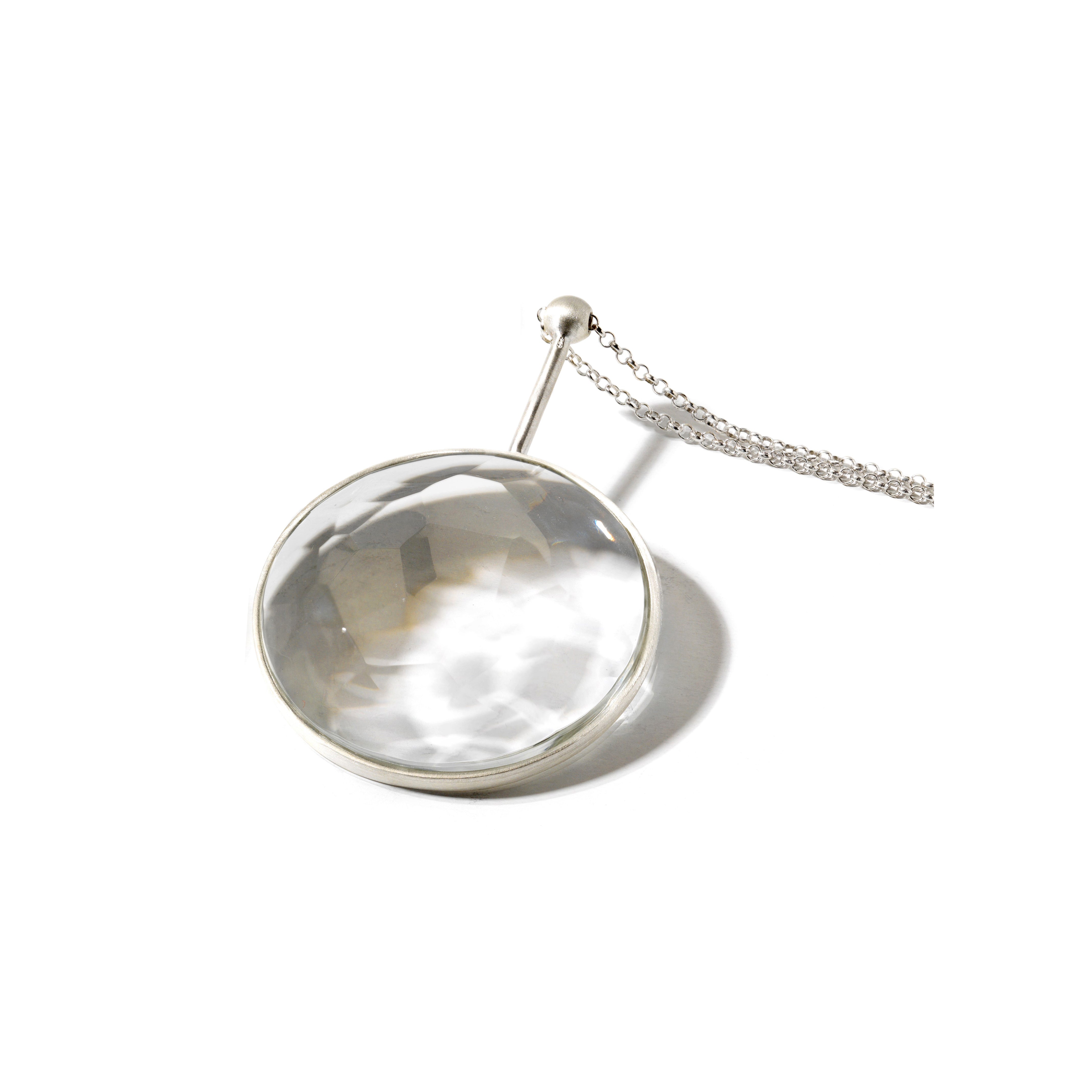 The Lobmeyr Crystal Jellies is a round magnifying glass with a shiny metal frame and handle, crafted using exquisite glass-cutting techniques. Its attached to a delicate chain and rests on a smooth white surface.