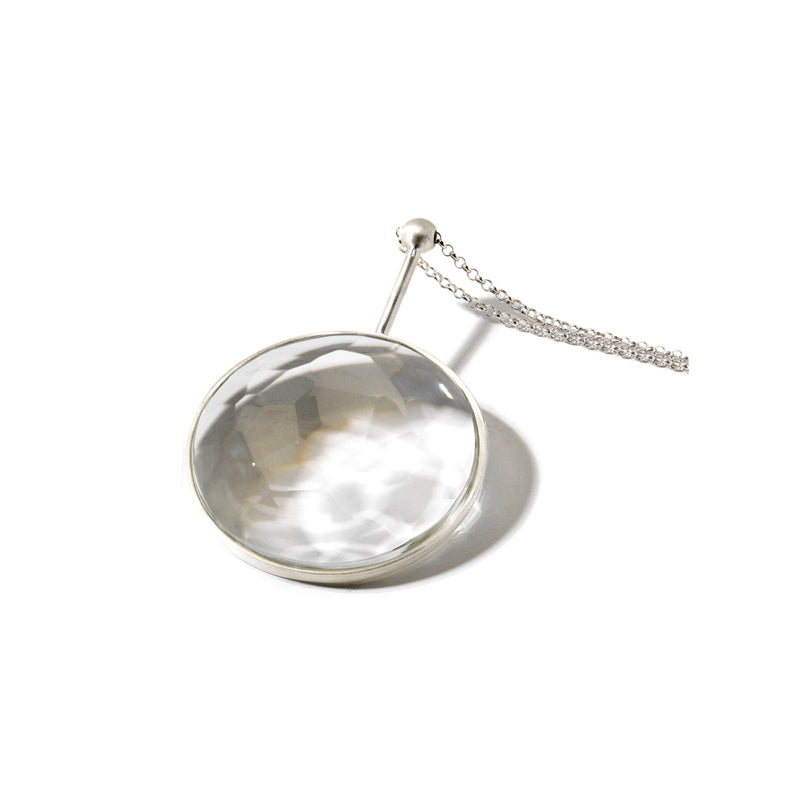 The Lobmeyr Crystal Jellies is a round magnifying glass with a shiny metal frame and handle, crafted using exquisite glass-cutting techniques. Its attached to a delicate chain and rests on a smooth white surface.