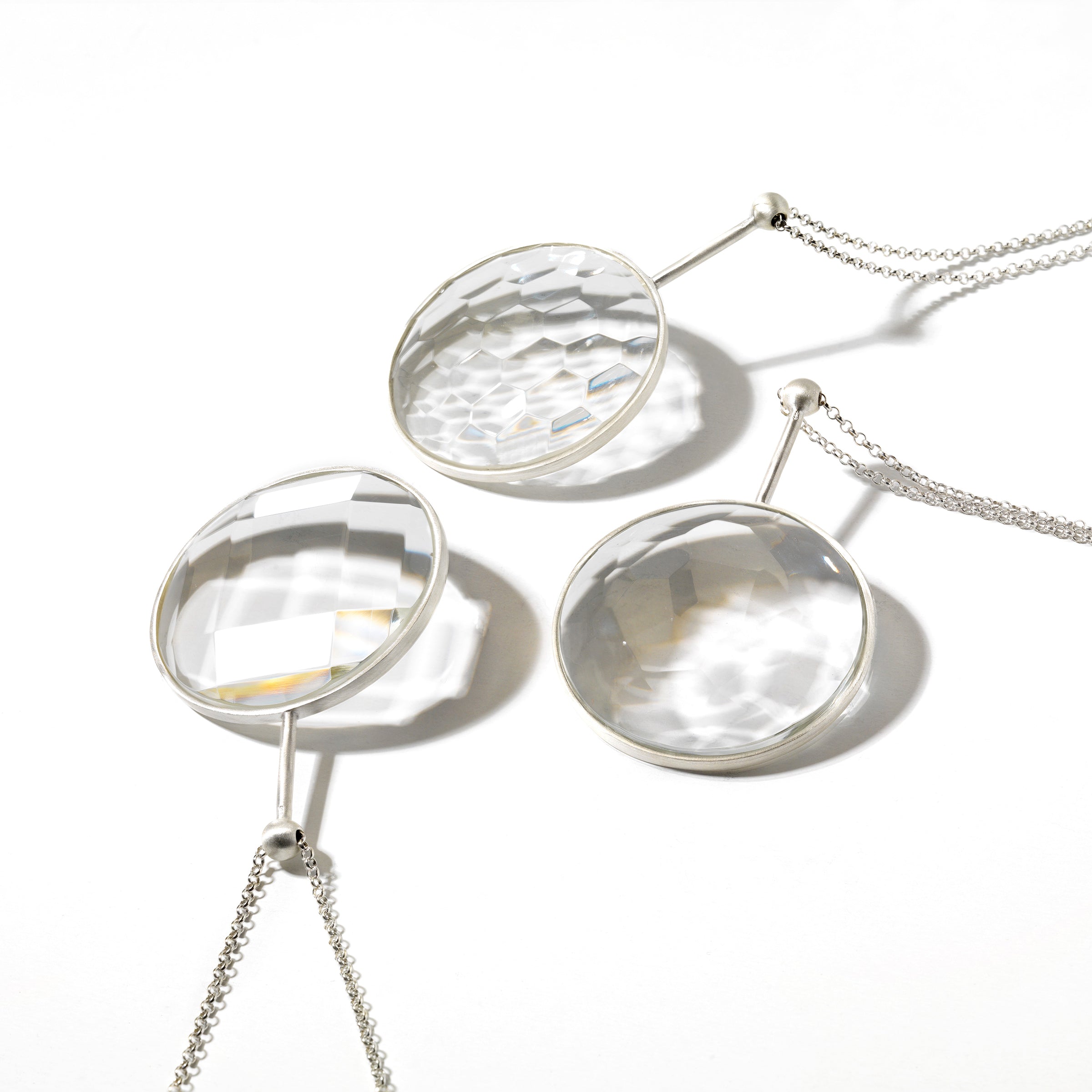Three Lobmeyr Crystal Jellies, featuring round faceted lead crystal pendants on silver chains, are displayed on a white surface. The crystals refract light into prismatic patterns, enhancing the smooth look with a textured effect.