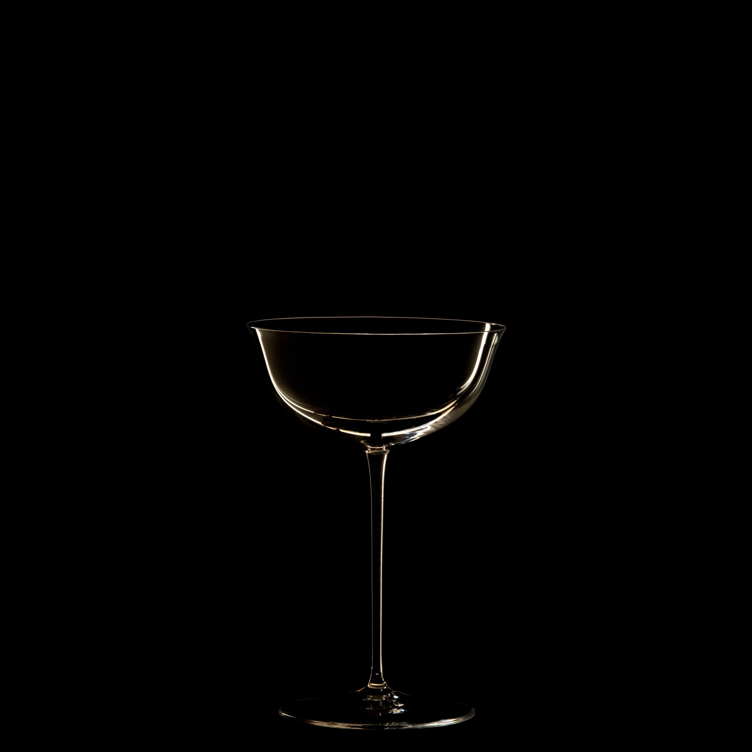 A single, elegant coupe glass from Lobmeyrs Drinking Set No. 238 - Patrician is dimly lit against a black background. Its silhouette reflects Josef Hoffmanns Wiener Werkstätte artistry, with its outline highlighting the exquisite shape and smooth curves.