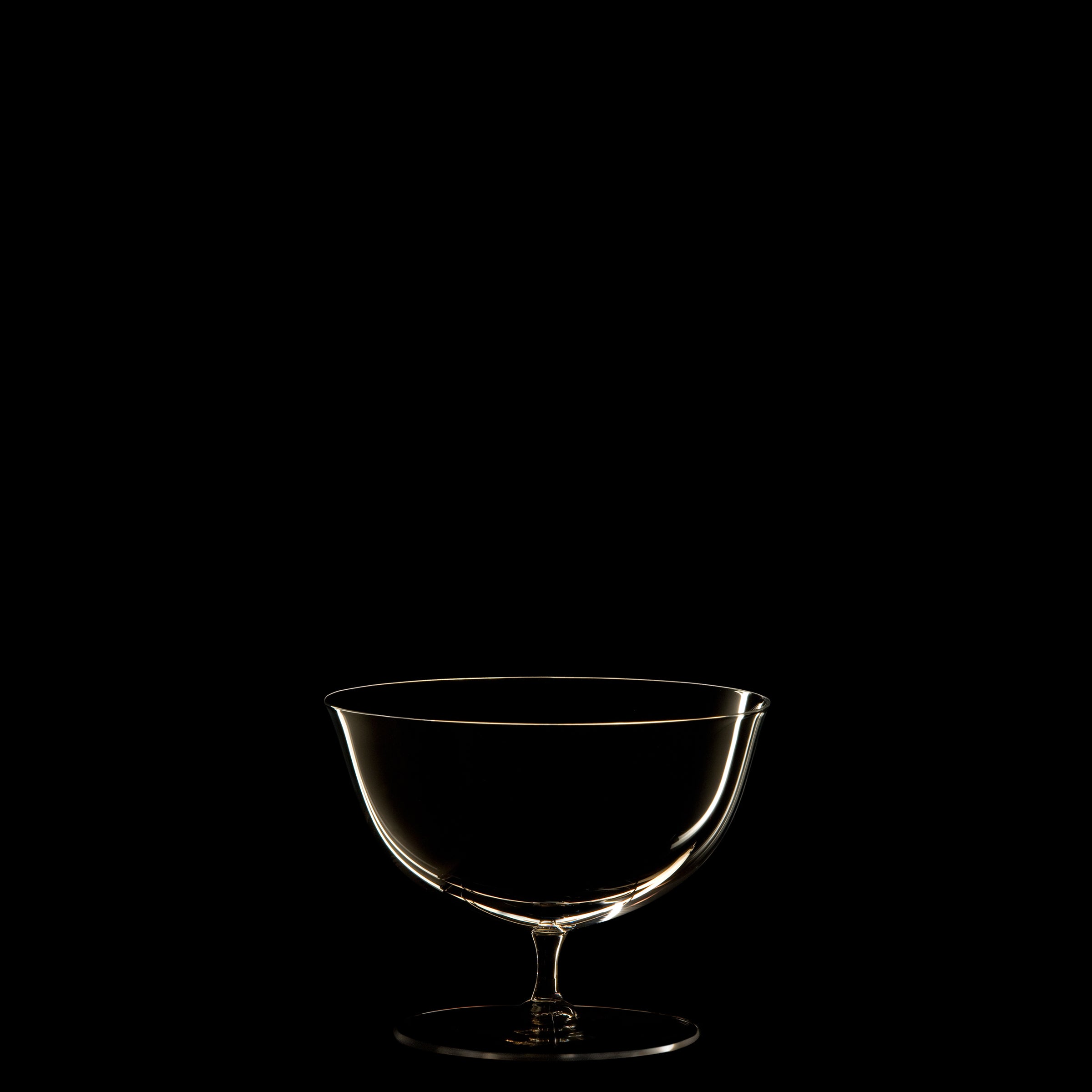 Set against a black background, a clear glass goblet from Lobmeyrs Drinking Set No. 238 - Patrician recalls Josef Hoffmanns mouth-blown crystal designs. Soft lighting accentuates its contours and delicate stem, capturing the dramatic elegance of the Wiener Werkstätte movement.