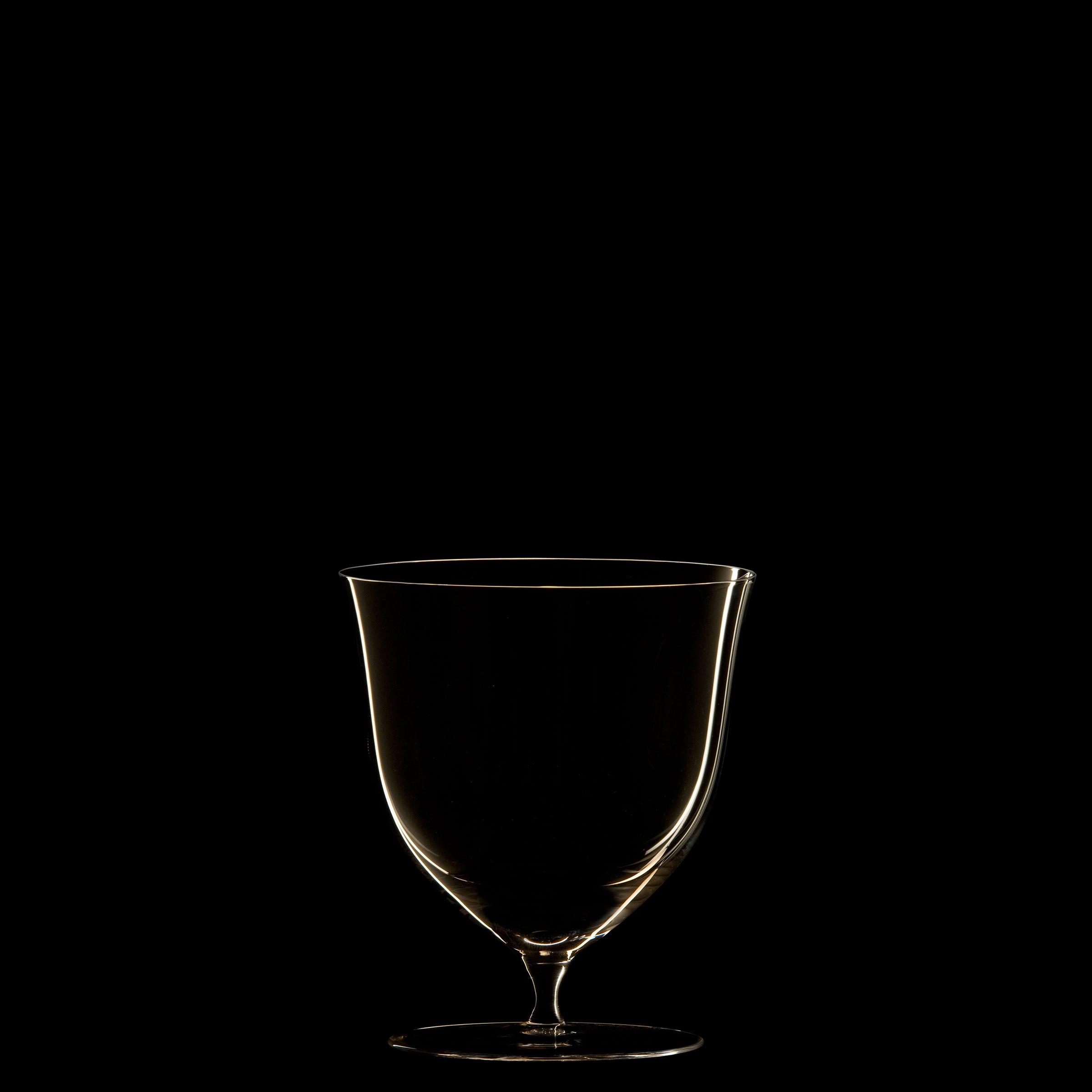 The Drinking Set No. 238 - Patrician by Lobmeyr features an elegant, mouth-blown crystal wine glass illuminated against a black background. Its empty form showcases a soft highlight along the rim and stem, exuding minimalist sophistication reminiscent of the Wiener Werkstätte style.