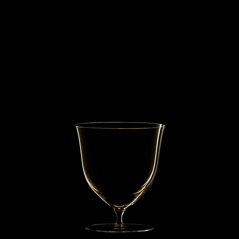 The Drinking Set No. 238 - Patrician by Lobmeyr features an elegant, mouth-blown crystal wine glass illuminated against a black background. Its empty form showcases a soft highlight along the rim and stem, exuding minimalist sophistication reminiscent of the Wiener Werkstätte style.