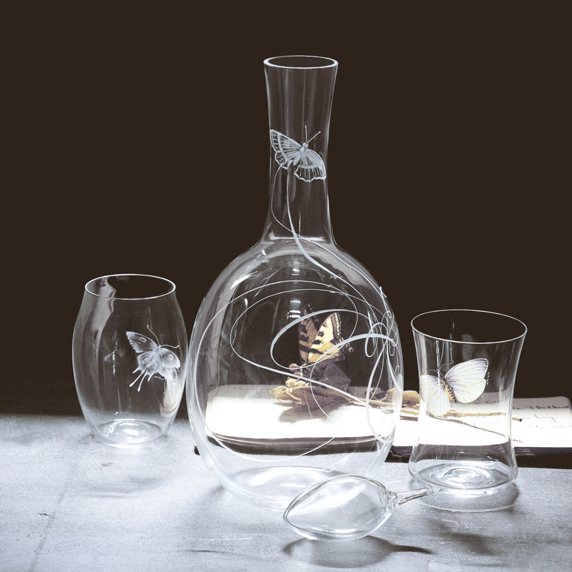 The Drinking Set No. 279 - Balloon by Lobmeyr features a mouth-blown glass decanter and two glasses, etched with Ted Muehlings butterfly designs. Set on a softly lit surface, the lead-free crystal captures light, highlighting whimsical patterns and casting subtle shadows.