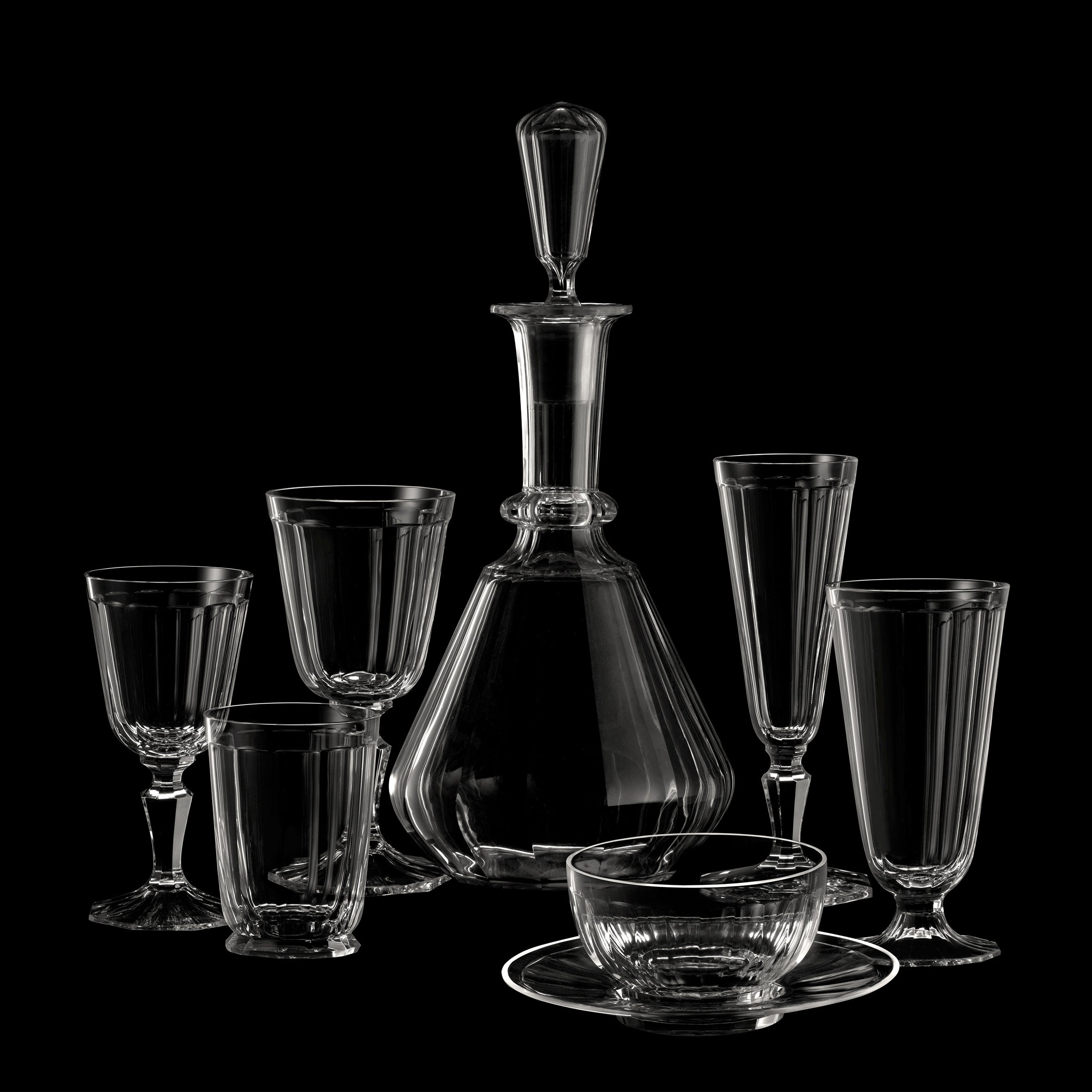A display of Lobmeyrs Drinking Set No. 98 - Palais with Facette Cut on a black background features a central decanter, six diverse glasses, and a teacup with saucer, emphasizing the transparency and intricate design of Ludwig Lobmeyrs mouth-blown crystal.