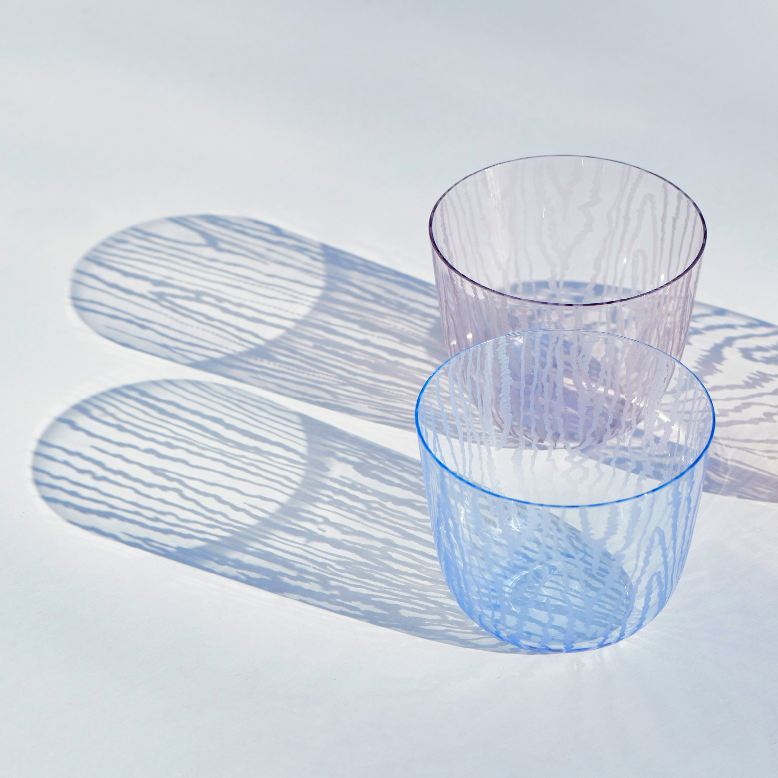 Two Lobmeyr Moiré Water Tumblers, one purple and one blue, rest on a white surface. Light creates striped shadows through the lead-free crystal, forming circular patterns reminiscent of a Moiré pattern.