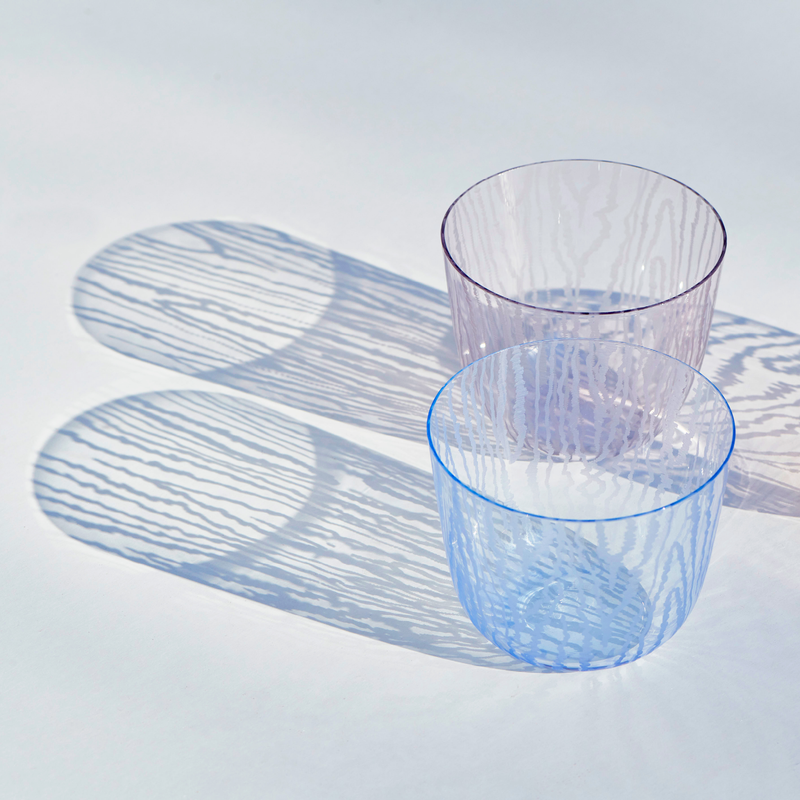 Two Lobmeyr Moiré Water Tumblers, one purple and one blue, rest on a white surface. Light creates striped shadows through the lead-free crystal, forming circular patterns reminiscent of a Moiré pattern.