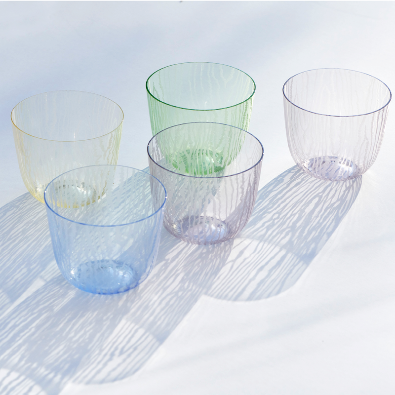 Five Lobmeyr Moiré Water Tumblers in pastel shades of green, yellow, blue, pink, and purple are arranged on a white surface. They create intricate moiré patterns with their shadows, producing a delicate design.