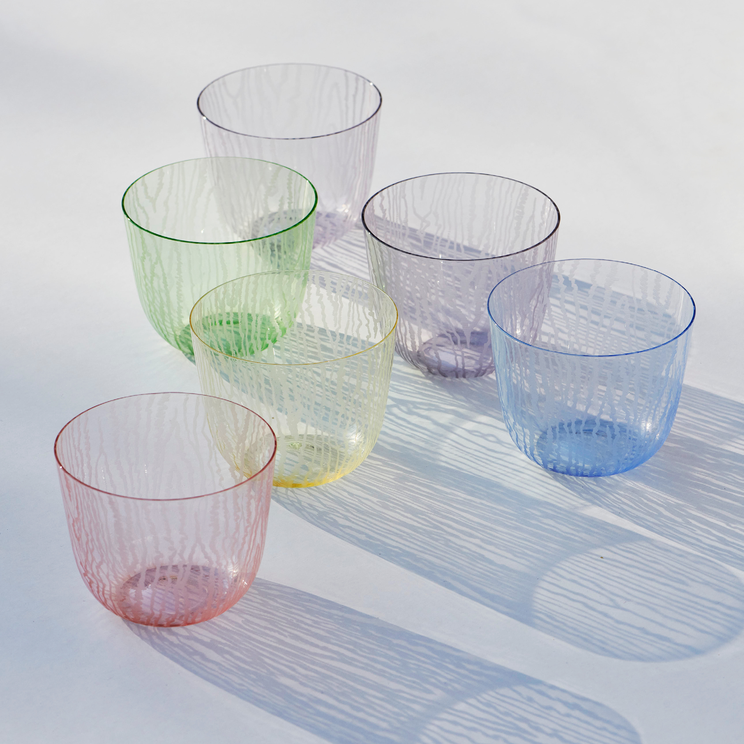 Six Lobmeyr Moiré Water Tumblers, translucent and textured, in pastel hues (green, pink, yellow, blue, purple), are set on a white surface. Sunlight casts intricate shadows through them, highlighting their moiré patterns and creating a visually appealing display.