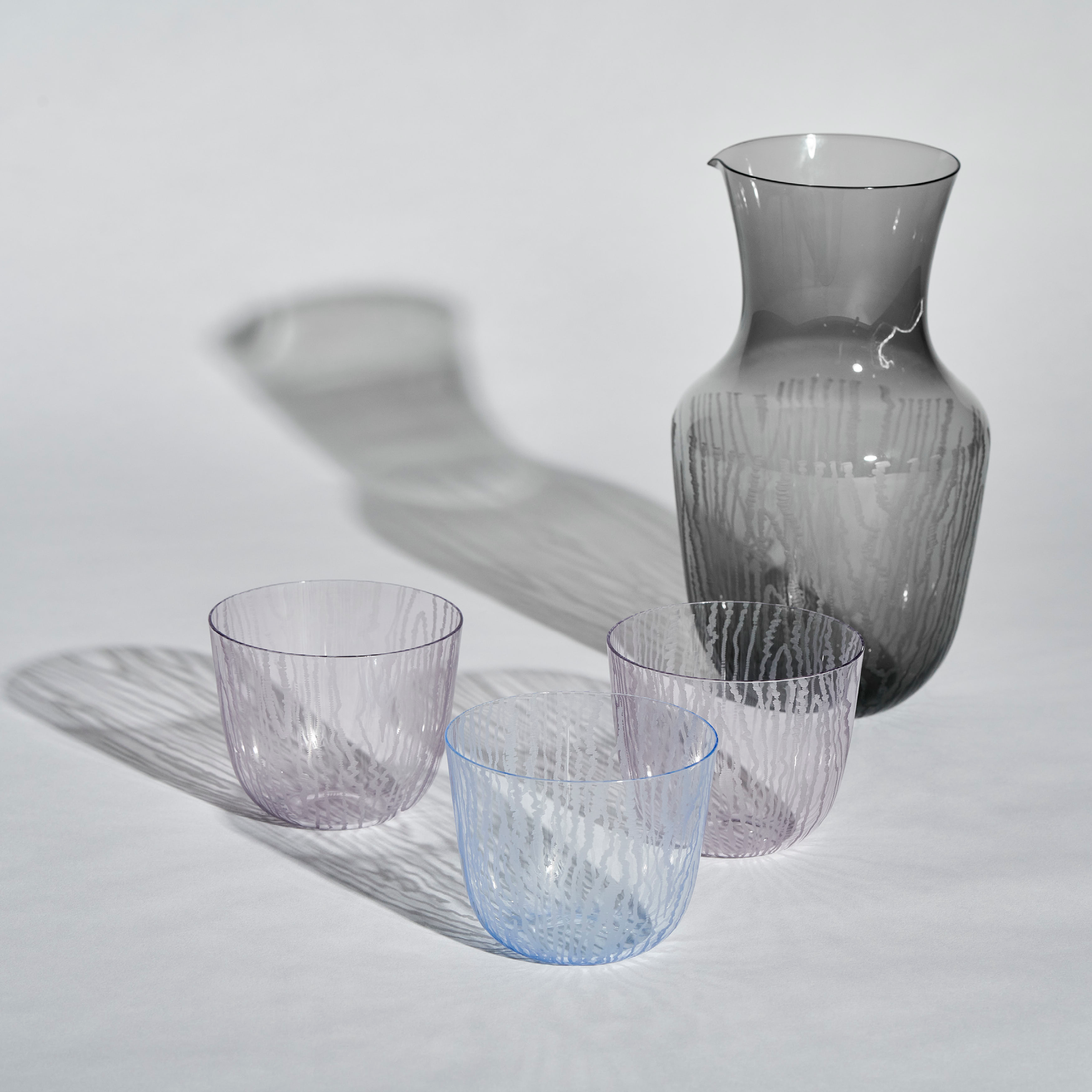 A Lobmeyr Moiré Water Pitcher in gray glass and three transparent textured glasses in light purple and blue are arranged on a surface, with lighting creating distinct shadows that highlight their intricate moiré pattern.