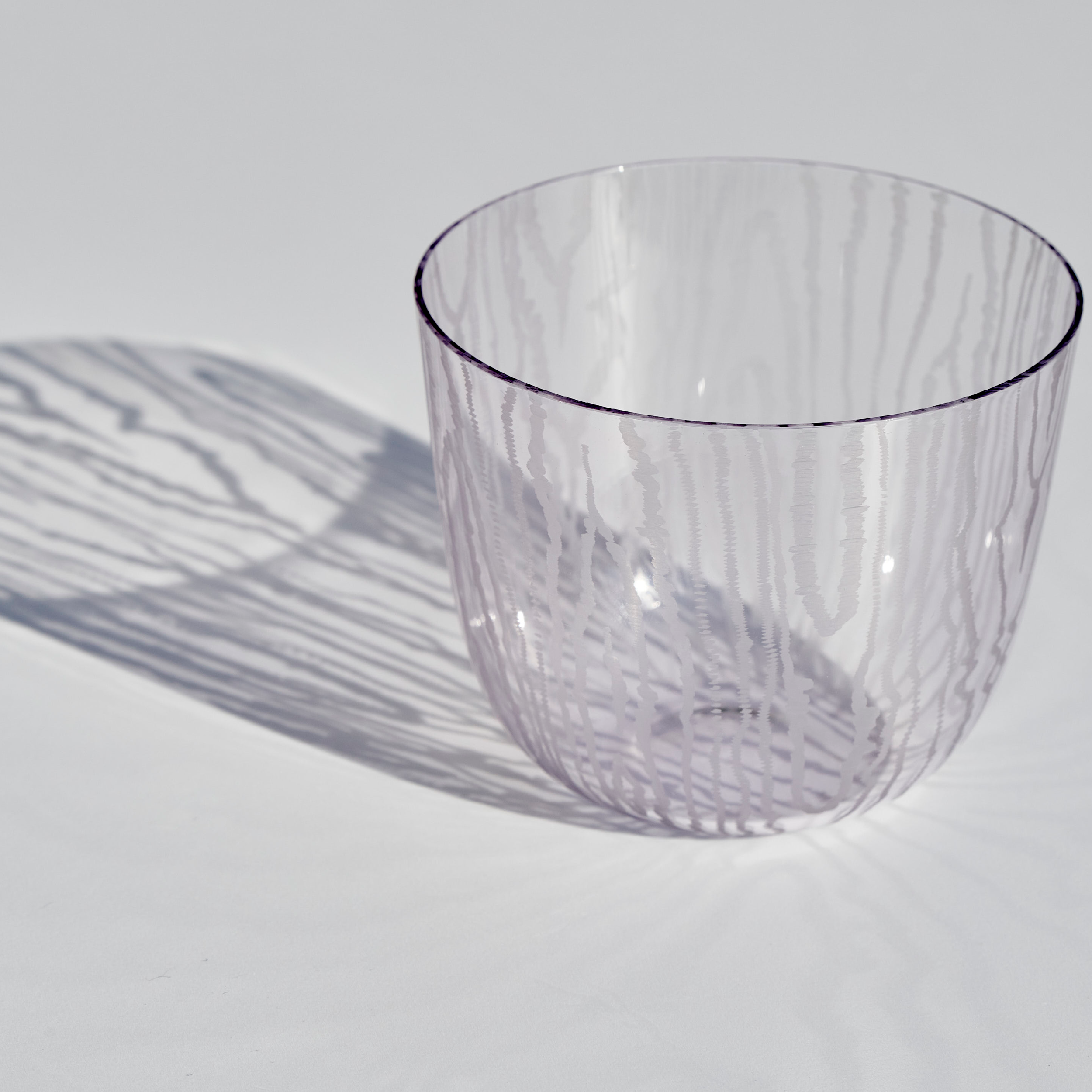 The Lobmeyr Moiré Water Tumbler by Lobmeyr, crafted from lead-free crystal with a wavy texture, casts intricate shadows on a light gray background that mirror the tumblers design and create a Moiré pattern.