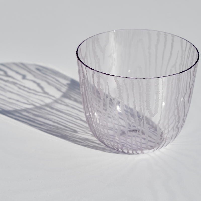 The Lobmeyr Moiré Water Tumbler by Lobmeyr, crafted from lead-free crystal with a wavy texture, casts intricate shadows on a light gray background that mirror the tumblers design and create a Moiré pattern.