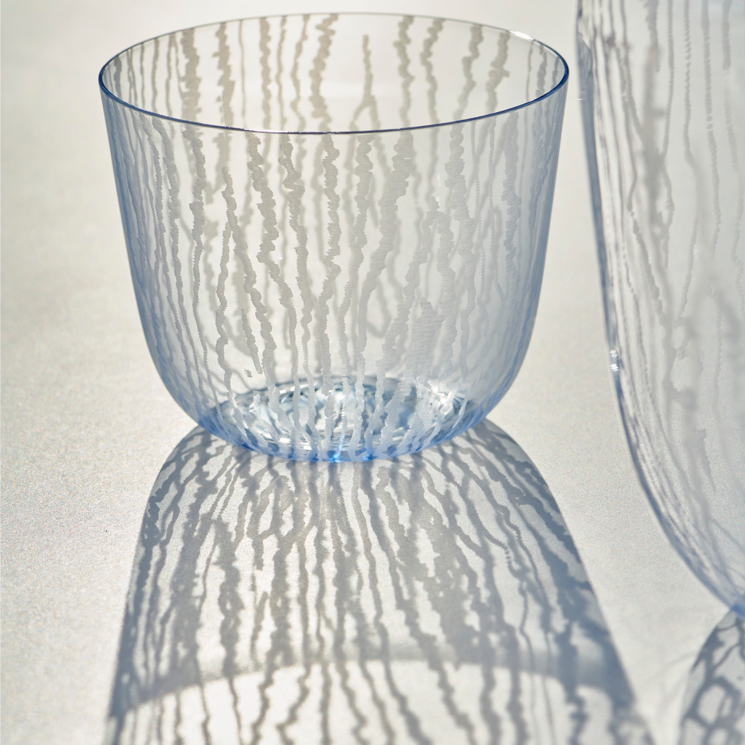 The Lobmeyr Moiré Water Tumbler, featuring translucent blue glass from the Alpha series, casts intricate shadows with its striped patterns reminiscent of a Moiré design. A partial view of a similar tumbler appears on the right, while lighting enhances its elegant textures and artistic effect.