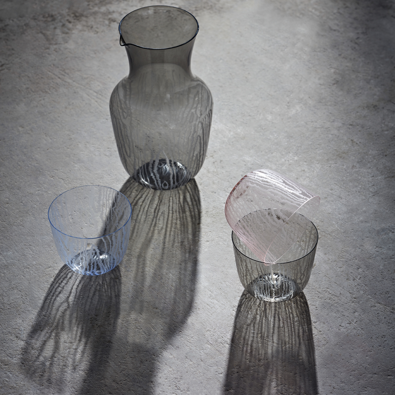 A Lobmeyr Moiré Water Pitcher and three textured, lead-free crystal glasses by Bodo Sperlein cast intricate shadows on a grey surface. One glass tips over, balancing on another upright glass, while the pitcher stands slightly apart.