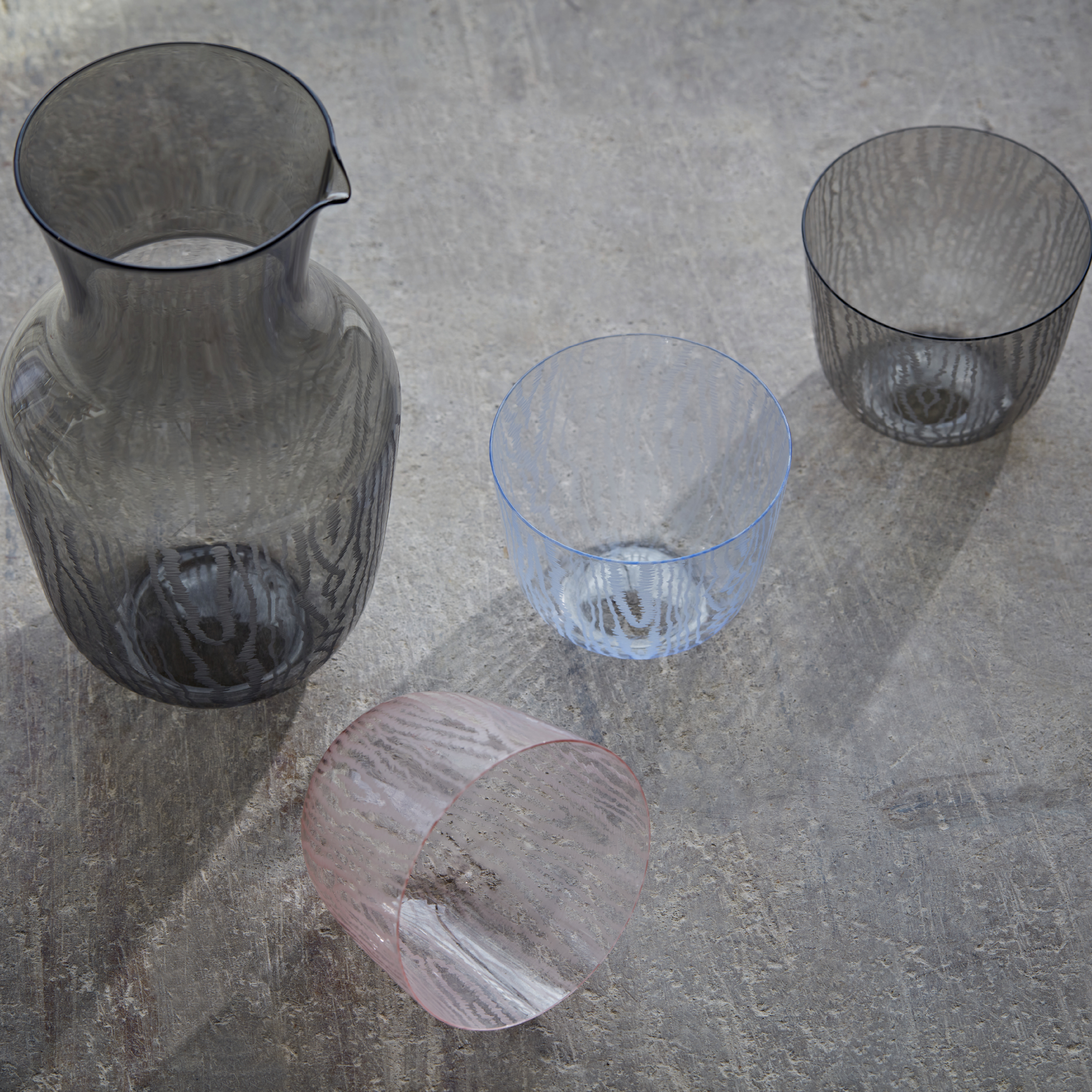 A Lobmeyr Water Pitcher by Bodo Sperlein and three textured, lead-free crystal glasses in black, blue, and pink are on a textured gray surface with soft shadows. The pink glass has a moiré pattern and is tilted on its side.