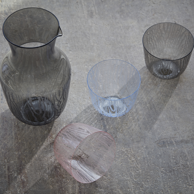 A Lobmeyr Water Pitcher by Bodo Sperlein and three textured, lead-free crystal glasses in black, blue, and pink are on a textured gray surface with soft shadows. The pink glass has a moiré pattern and is tilted on its side.