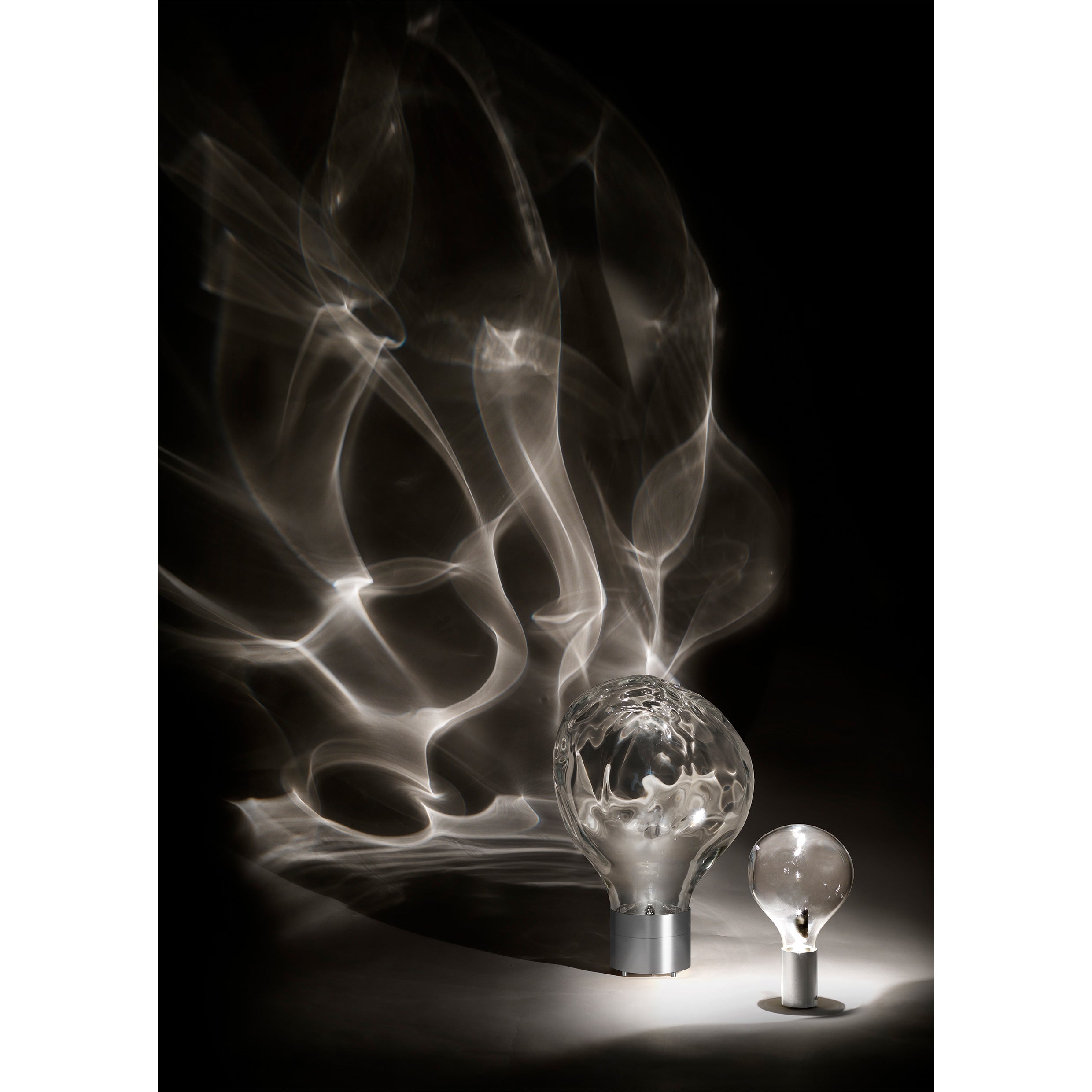 Against a dark background, two light bulbs cast their glow. The larger, Ripple Light by Lobmeyr, projects intricate swirling patterns that create an ethereal effect. Nearby, the smaller bulb of mouth-blown glass emits minimal light.