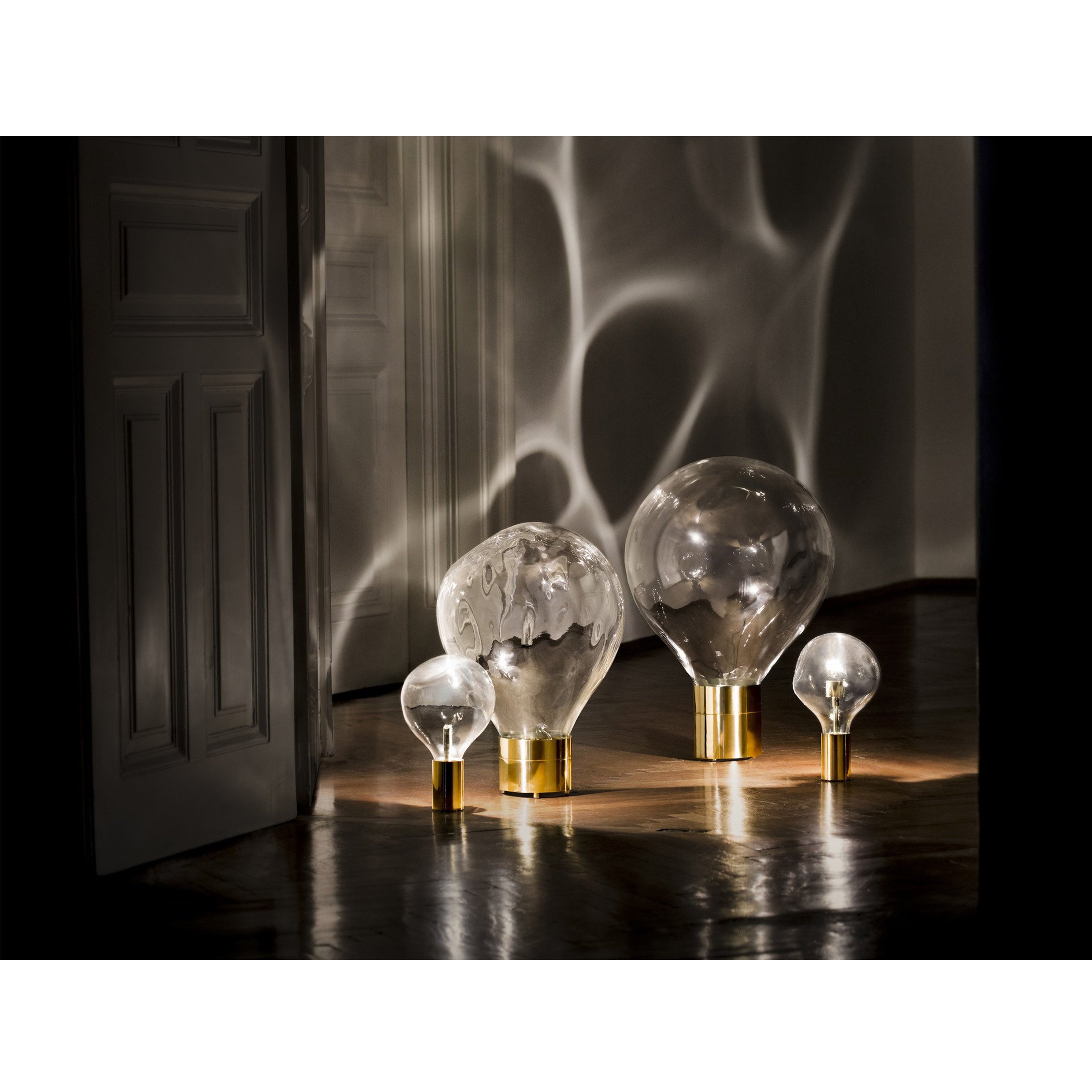 The dimly lit room features Ripple Light by Lobmeyr: four oversized, mouth-blown glass bulbs with gold bases rest on a polished wooden floor, casting soft reflections and ripple patterns on the walls and floor.