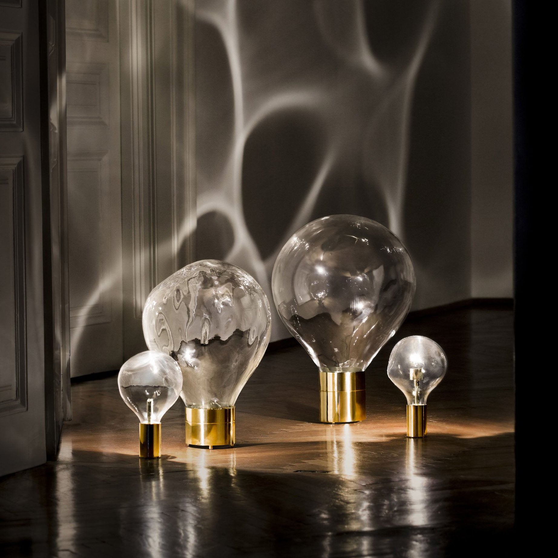 Four Ripple Light lamps by Lobmeyr, crafted from mouth-blown glass with gold bases, are set on a wooden floor. Their light paints intricate moving ripple patterns of shadows and reflections on the dark walls and floor, creating a moody and artistic atmosphere.