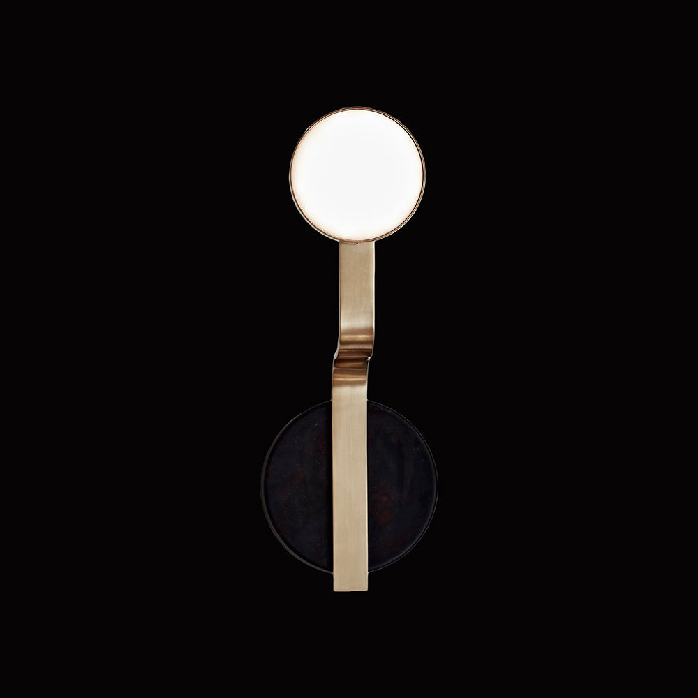 The Script Sconce by Lobmeyr is a modern minimalist wall light with a gold metallic frame, reminiscent of Viennese Art Deco. It features a round white disc at the top and a contrasting black circle below, creating an elegant design against a black background.