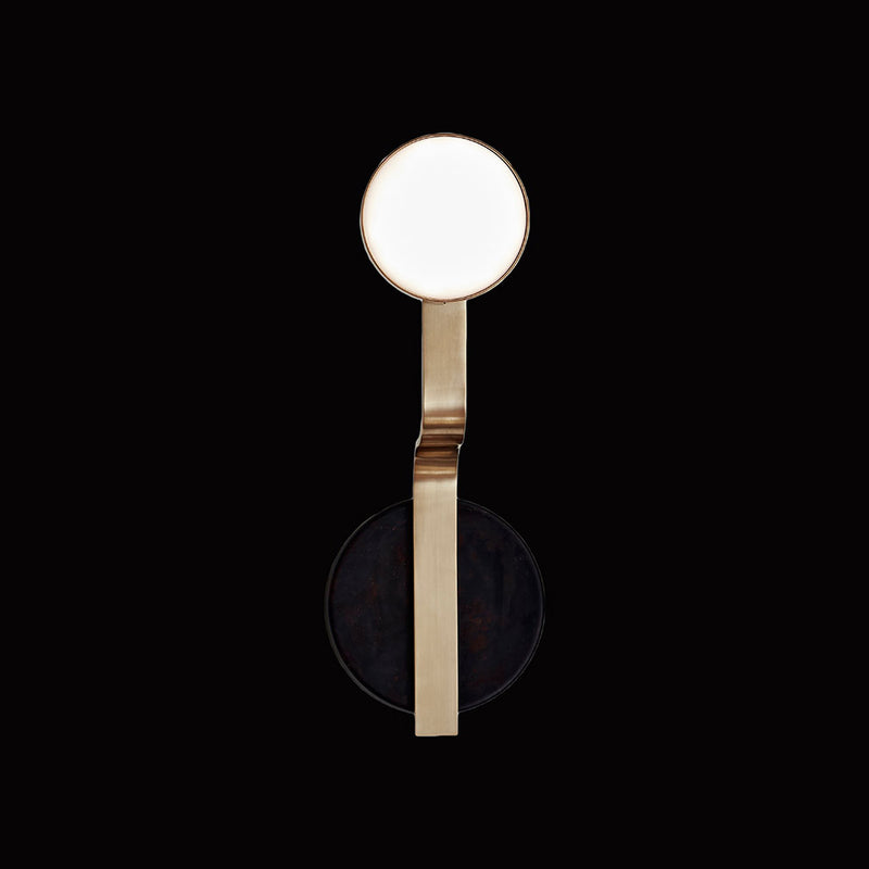 The Script Sconce by Lobmeyr is a modern minimalist wall light with a gold metallic frame, reminiscent of Viennese Art Deco. It features a round white disc at the top and a contrasting black circle below, creating an elegant design against a black background.
