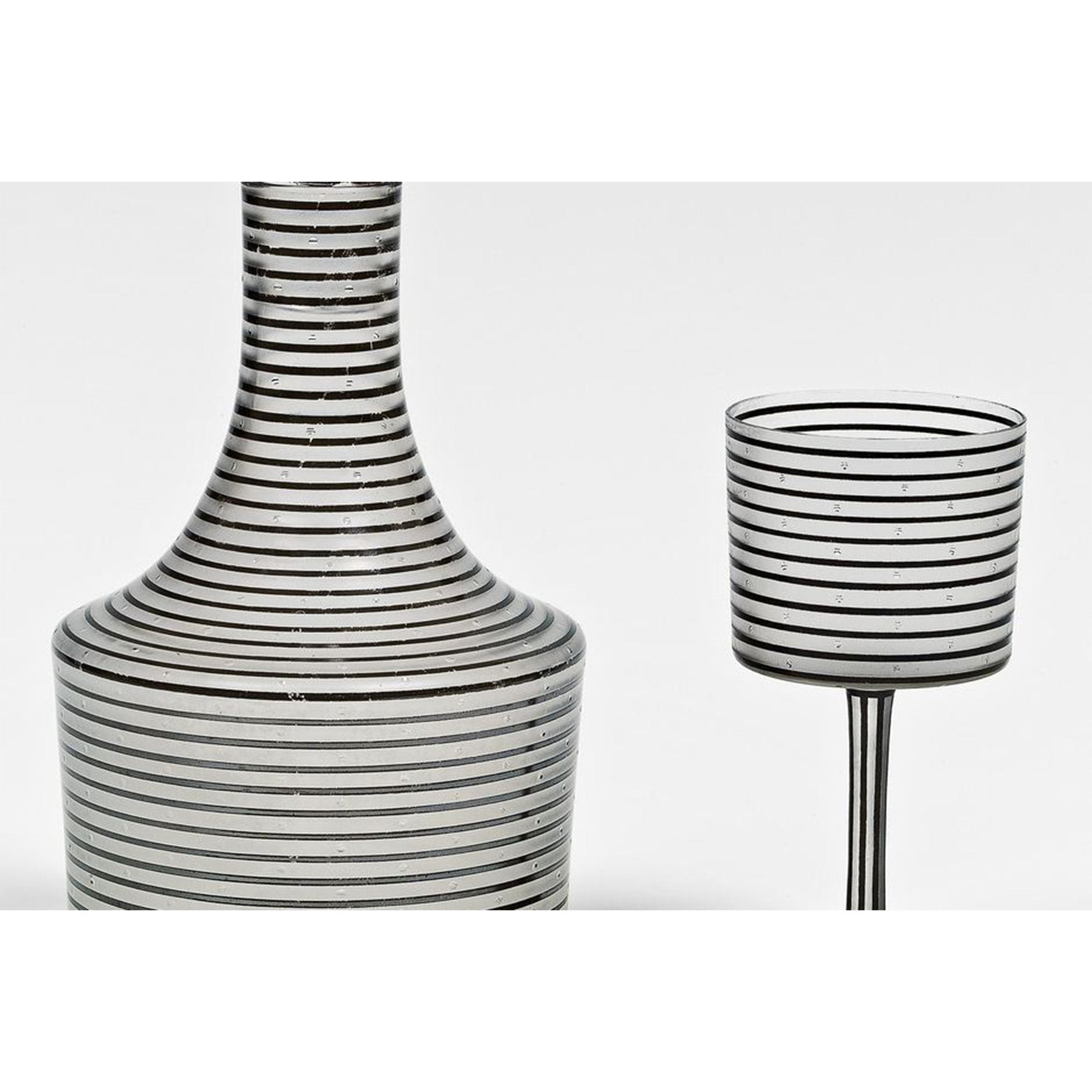 The Drinking Set Serie A by Lobmeyr includes a mouth-blown crystal decanter and matching glass, featuring a vertical black-lined pattern on white. The decanter has a wide base with a narrow neck, while the glass rests on an elegant slender stem.