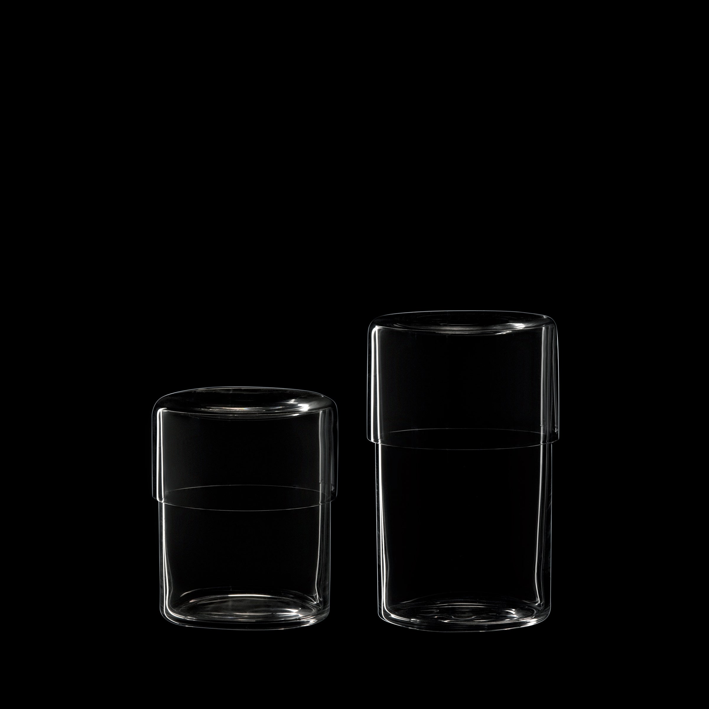 Two Strnad Box - Cigarette Boxes by Lobmeyr, crafted from mouth-blown lead-free crystal with flat lids—one taller than the other—are showcased against a black backdrop, highlighting their minimalist cylindrical design that echoes Oskar Strnad and Lobmeyrs refined aesthetics.