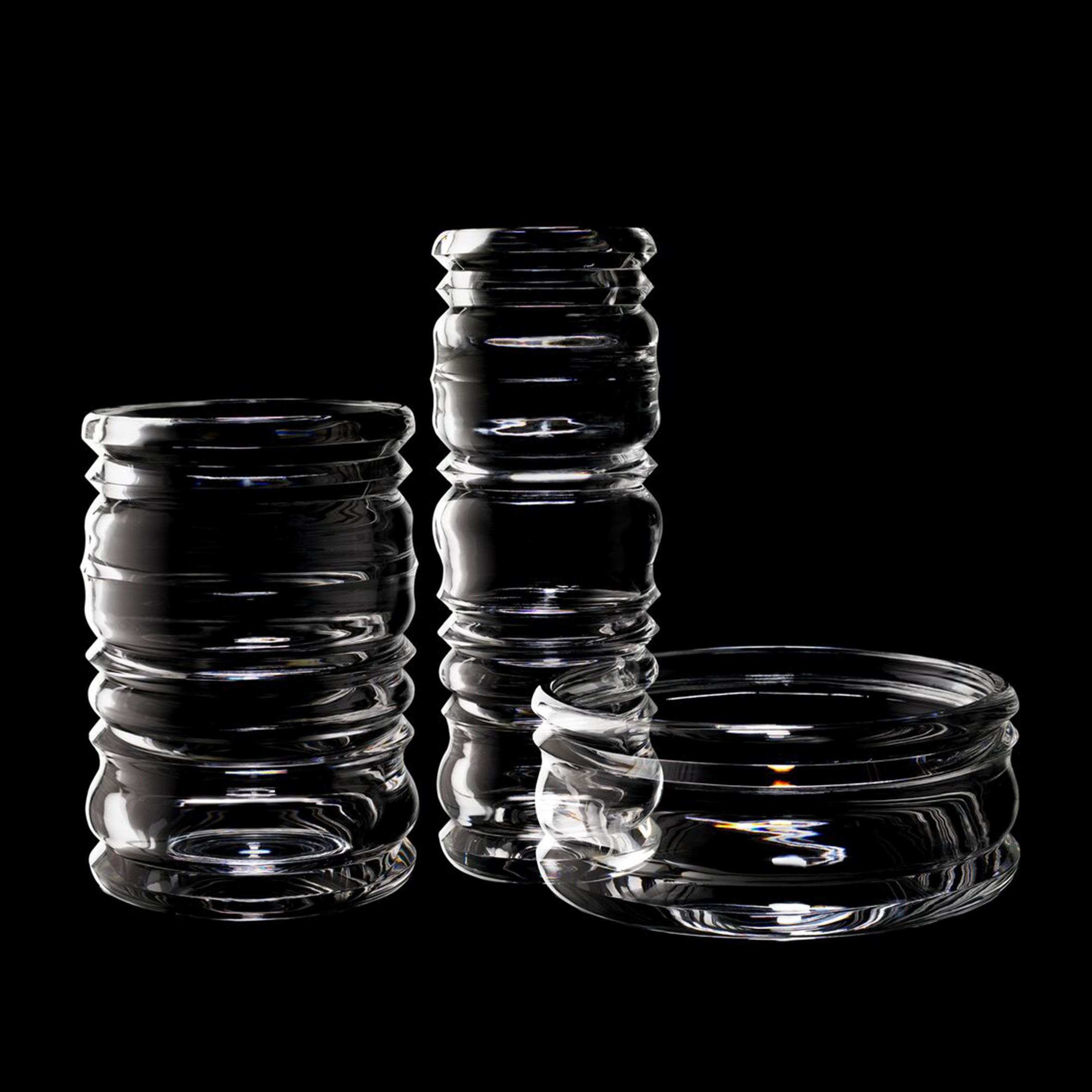 The Thom Series by Lobmeyr, designed by Sebastian Menschhorn, is showcased against a black backdrop. This stunning collection features two tall jars and one shallow bowl made of mouth-blown glass with ribbed designs, all in an exquisite, clear finish.