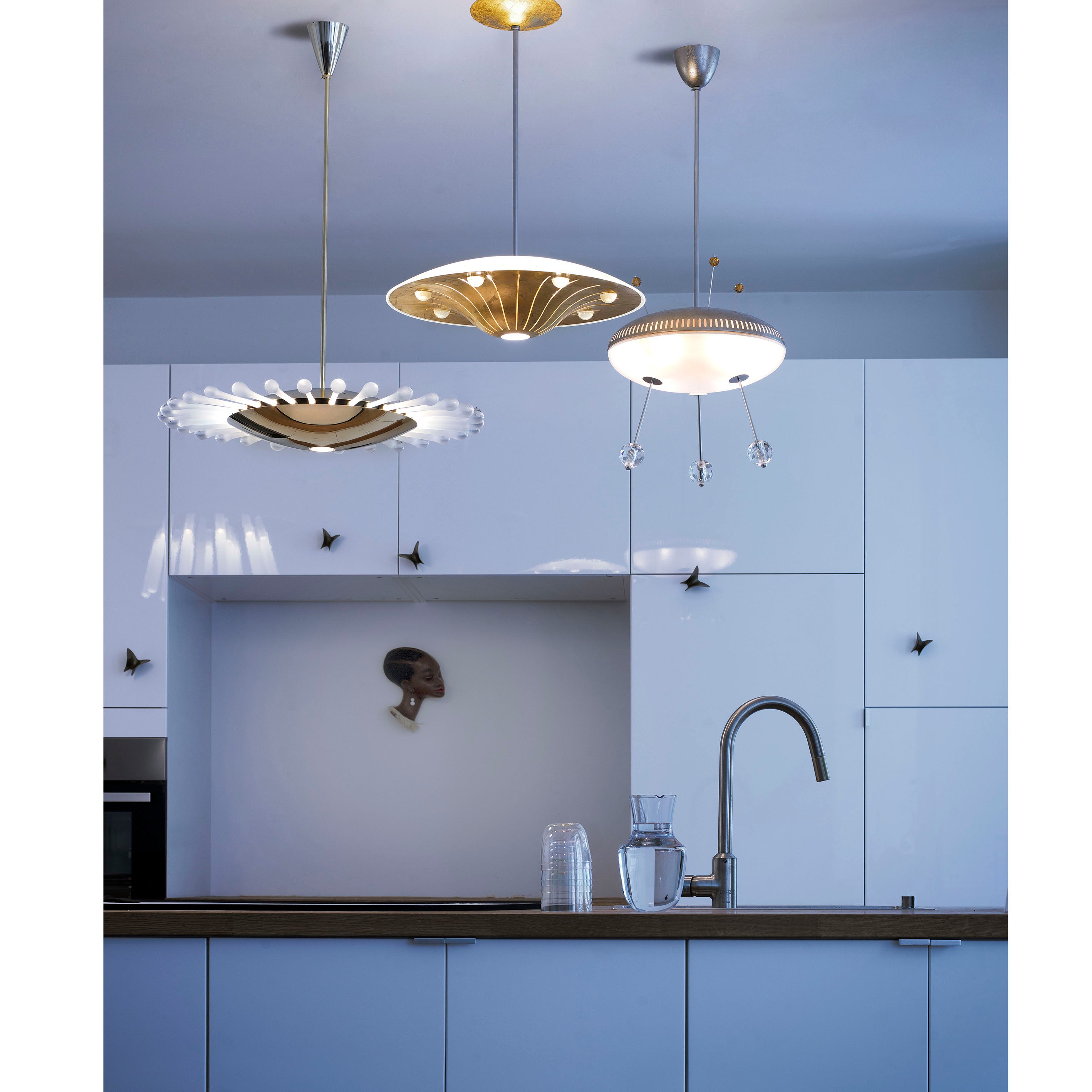 A modern kitchen with white cabinets and a wooden countertop, illuminated by three stylish Lobmeyr pendant lights. A water carafe and glass sit on the counter, while a Space Ship Ellen decorative face intriguingly gazes from the cabinet wall like something out of a UFO series.