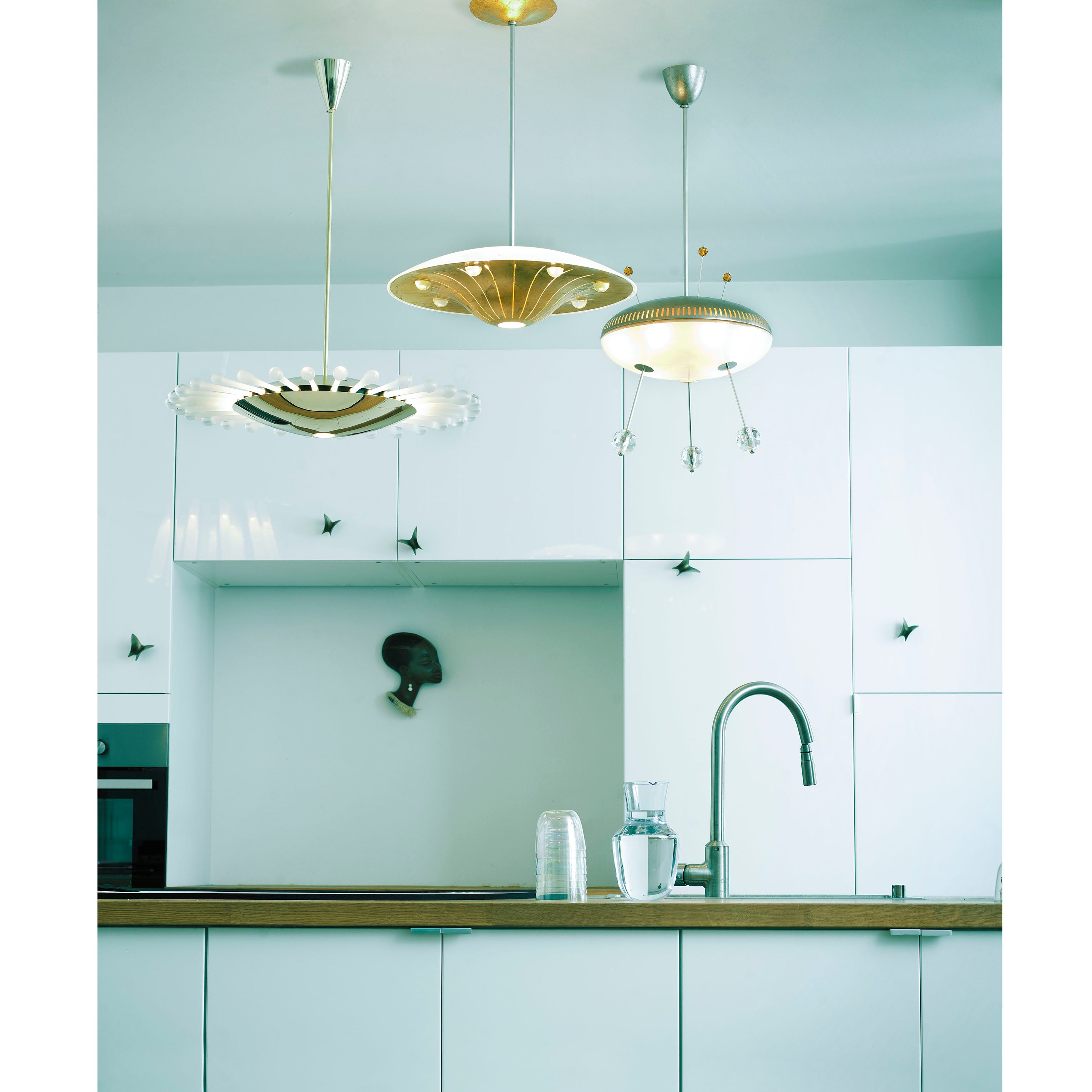 A modern kitchen with white cabinets and a wooden countertop features three unique Lobmeyr Space Ship Nyota pendant lights. Theres a curved faucet, a glass and carafe on the counter, and silhouette artwork on the wall.