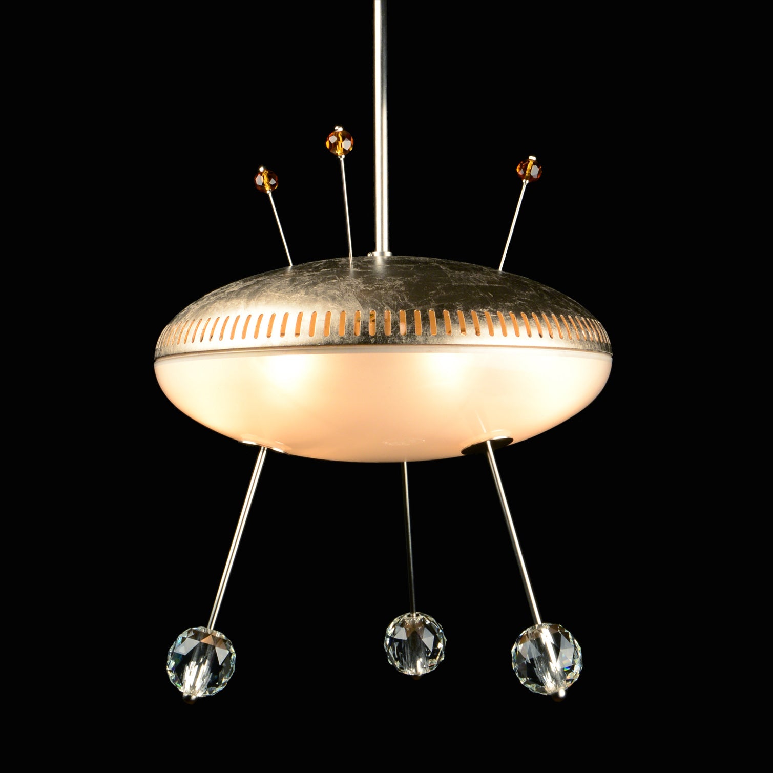 The Space Ship Orson by Lobmeyr features a UFO-shaped design with a metallic top and frosted glass underside, adorned with crystal-like accents hanging from thin rods amidst a black backdrop, reflecting space-age modernity.