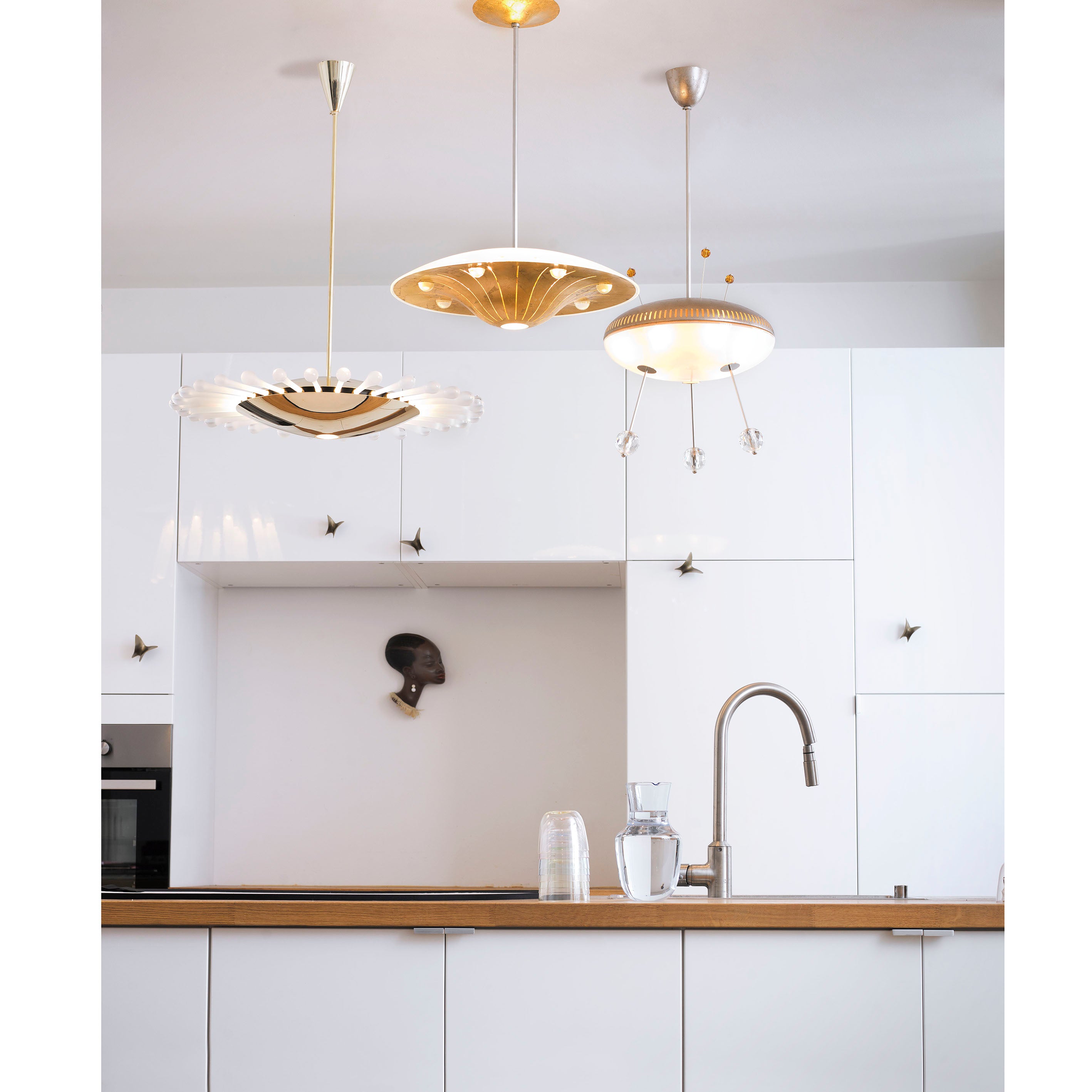 A modern kitchen features white cabinets and a wooden countertop. Three Lobmeyr Space Ship Orson pendant lights provide a space-age touch. A silver faucet sits next to empty glasses and a jug, while a small artwork of a persons head decorates the wall.