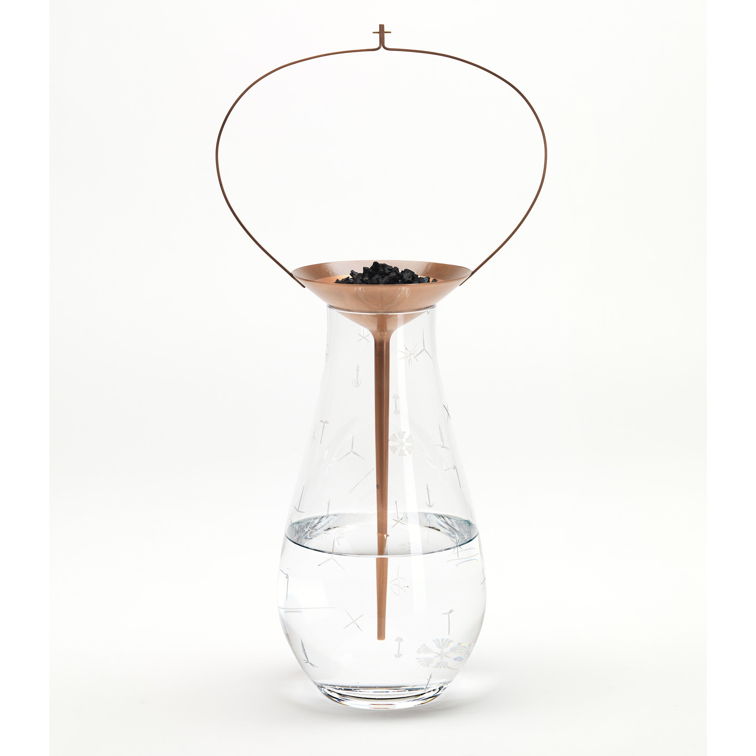 The Still Series by Lobmeyr features a glass container like a crystal vessel with clear liquid, topped by a copper-colored, cone-shaped strainer holding small black pieces and an elegantly arched thin circular wire handle.