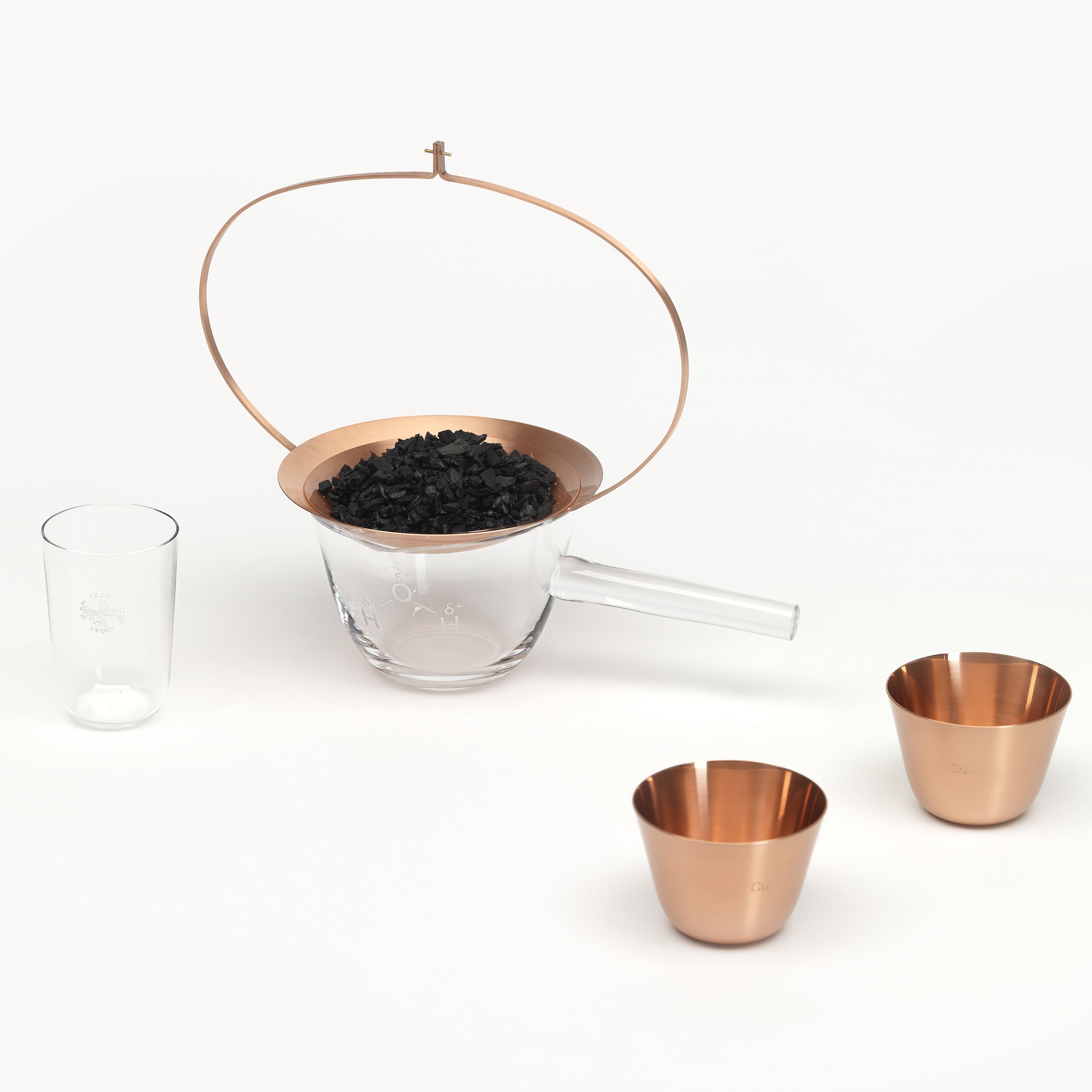The Lobmeyr Still Series features a glass container with a handle and copper strainer holding charcoal, complemented by two copper measuring cups and a crystal vessel, all elegantly placed on a pristine white surface.