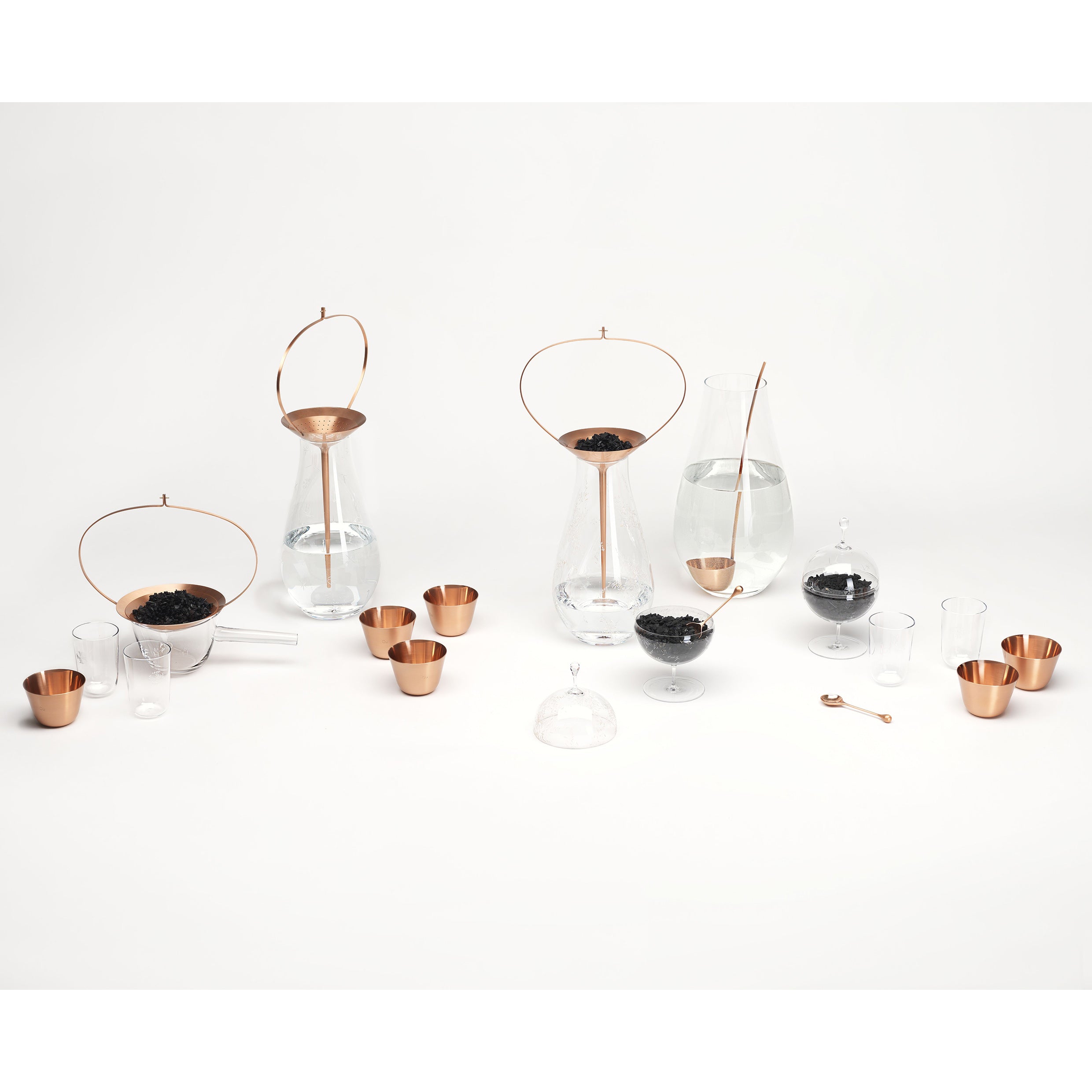 The Still Series by Lobmeyr features elegant glass decanters and copper cups with handles, some filled with black pebbles or liquid, displayed on a white surface. This minimalist set highlights a clean, modern aesthetic with natural materials and geometric shapes.