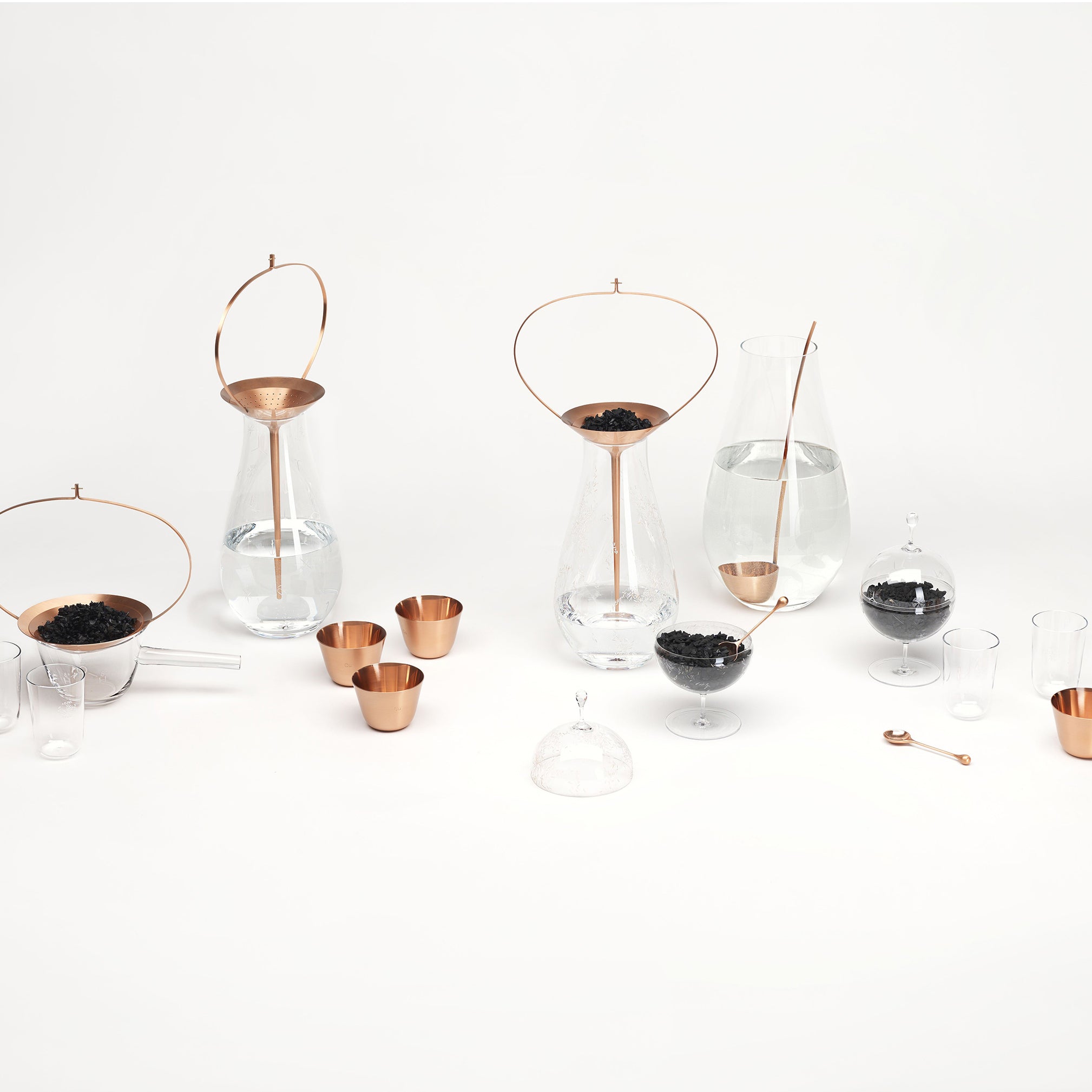 The Still Series by Lobmeyr features crystal vessels with copper accents, including carafes with tea strainers, small bowls, glasses, a mixing spoon, and loose leaf tea. Displayed on a white background for a minimalist and contemporary look.