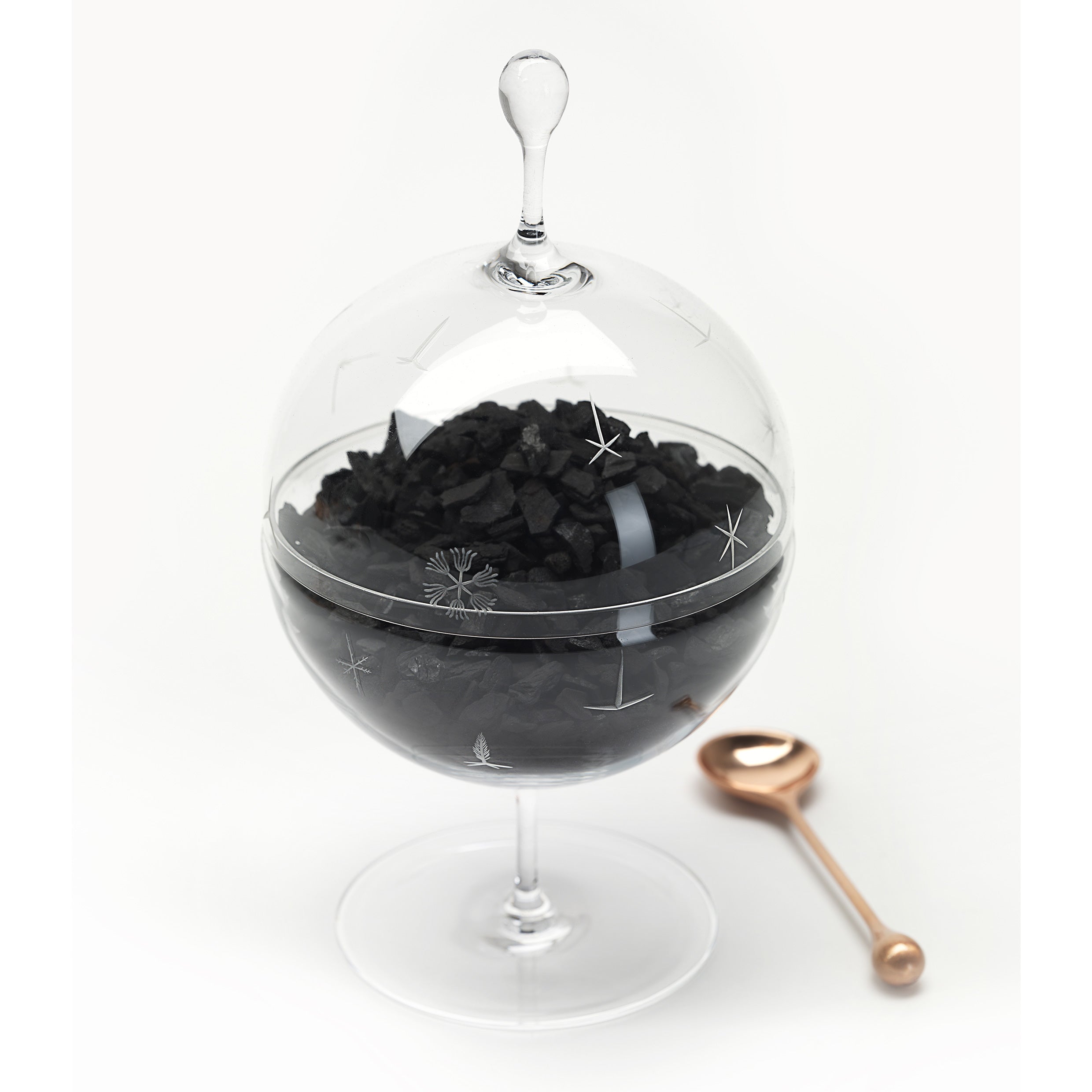 The Still Series by Lobmeyr is a transparent globe-shaped container on a stand, resembling crystal vessels. It is filled with black rock-like contents, adorned with star engravings and a top handle. Beside it rests a small copper spoon on the white surface, completing this intriguing tableau.