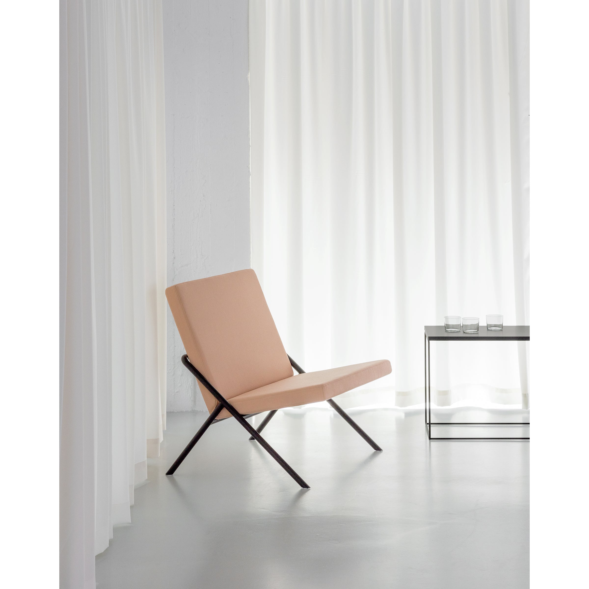 A minimalist room showcases the DL2 Euclides Lounge Chair by Loehr with pale peach upholstery and dark wooden legs, providing comfort near white curtains. A small glass table with two water glasses completes the elegant setup on a light gray floor.
