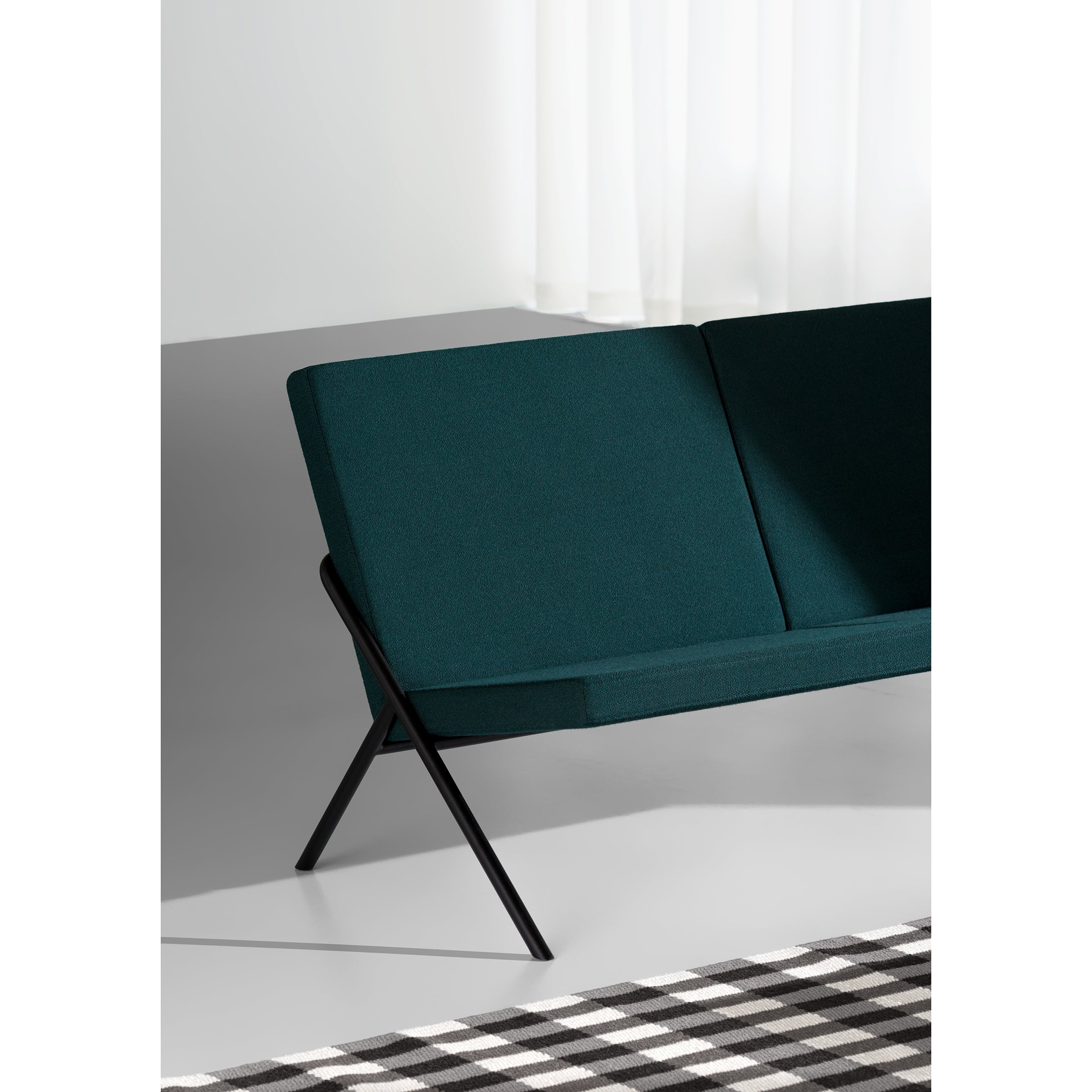 The DL2 Euclides Two-Seater by Loehr boasts a modern, sleek design with luxurious seating, featuring a deep green cushion and black metal frame. It sits in a minimal space with gray flooring, white curtains, and is partially enhanced by a black-and-white checkered carpet.