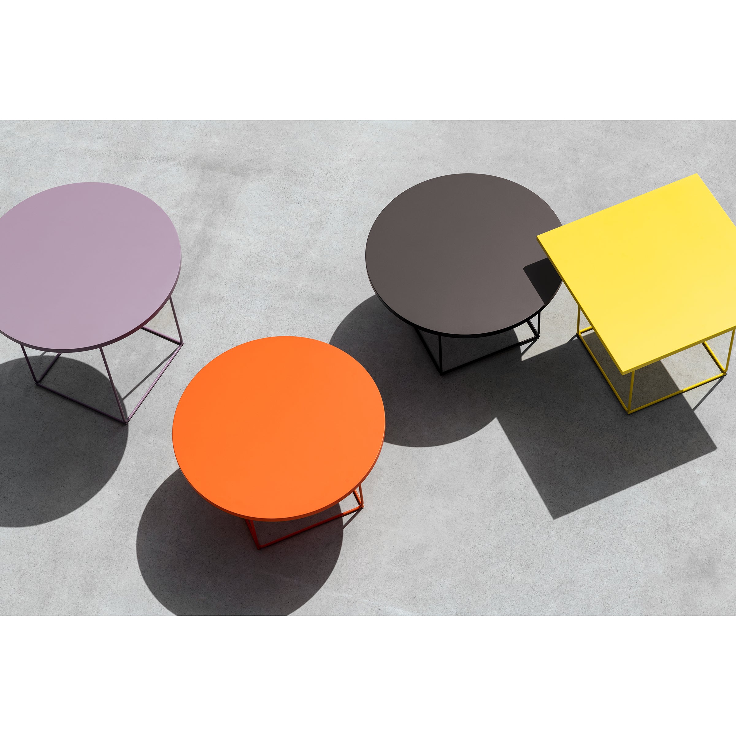 Four geometric tables rest on a concrete surface: three round coffee tables called the DL3 Umbra Table Round, available in vibrant purple, orange, and dark brown shades by Loehr, and a square yellow table. Their umbrae interact warmly, creating a modern minimalist aesthetic.