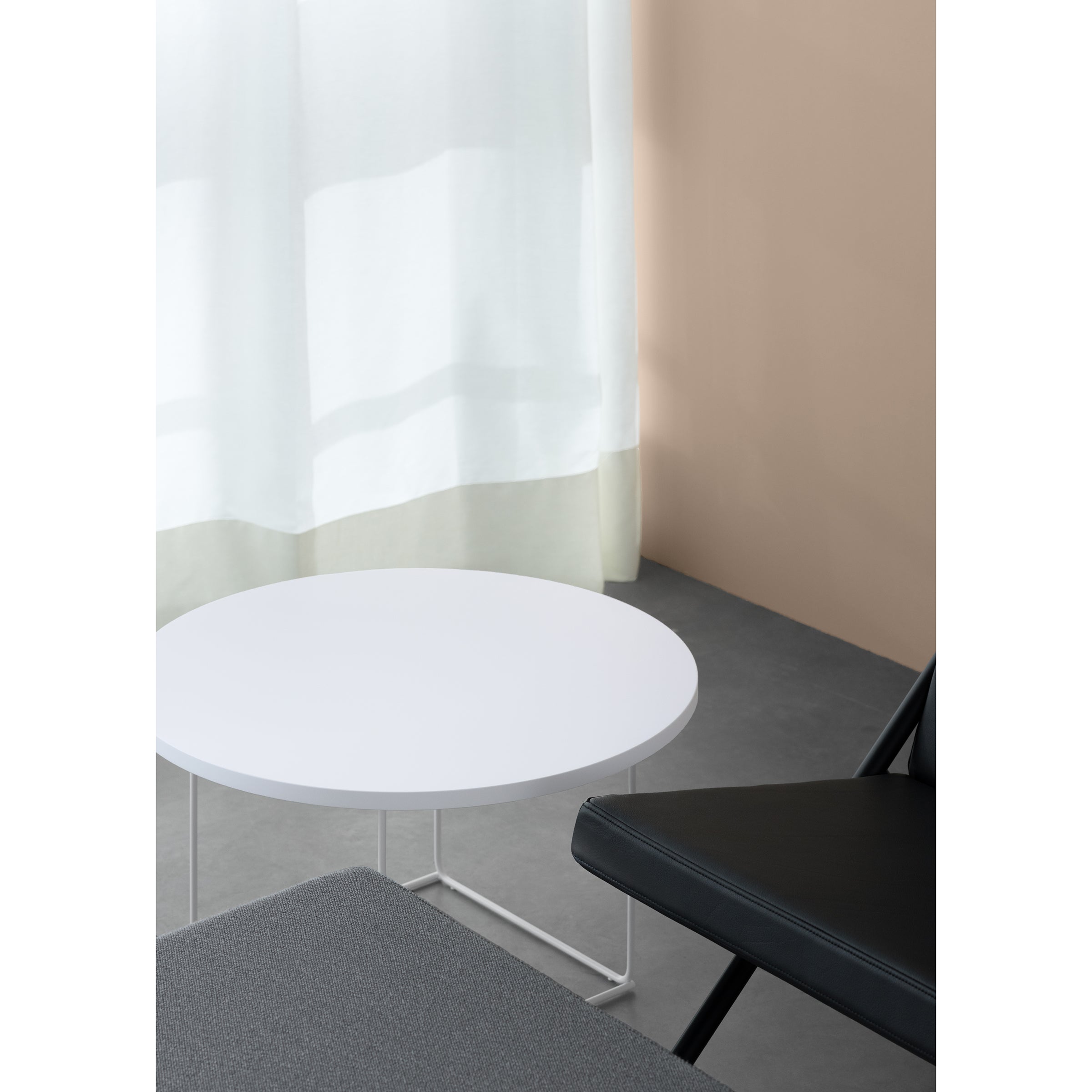 A minimalist room features a Loehr DL3 Umbra Table Round, a black chair, and a gray surface. A sheer white curtain drapes over a beige wall, letting in soft light.
