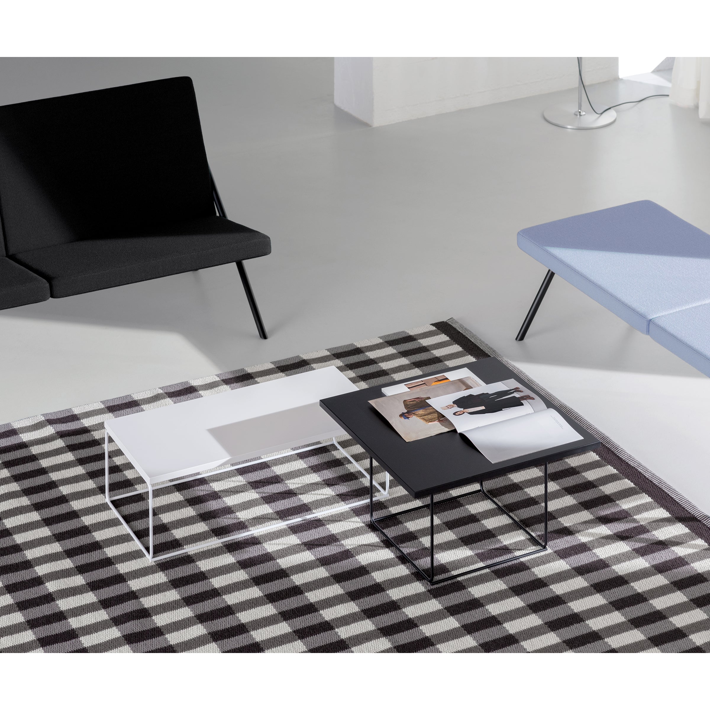 A minimalist living room features a black and white plaid rug, a round UMBRA coffee table, and two Loehr DL3 Umbra Table Squares, one black and one white, topped with magazines. A black sofa and light gray chair sit by large windows with sheer curtains, enhancing the modern design.