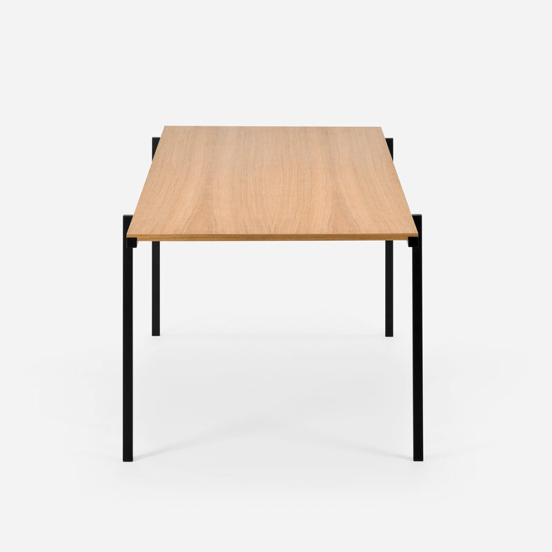 The DL5 Neo Table by Loehr, inspired by De Stijl, features a minimalist wooden top with a natural finish. It is supported by sleek black steel tube legs against a white backdrop, highlighting its clean, modern rectangular design.