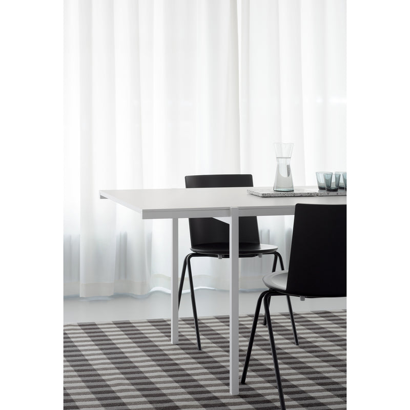Minimalist dining area with a DL5 Neo Table by Loehr and two black chairs on a checkered gray rug. A glass carafe and two cups sit on the table, showcasing De Stijl-inspired clean lines. Sheer white curtains provide a soft backdrop.