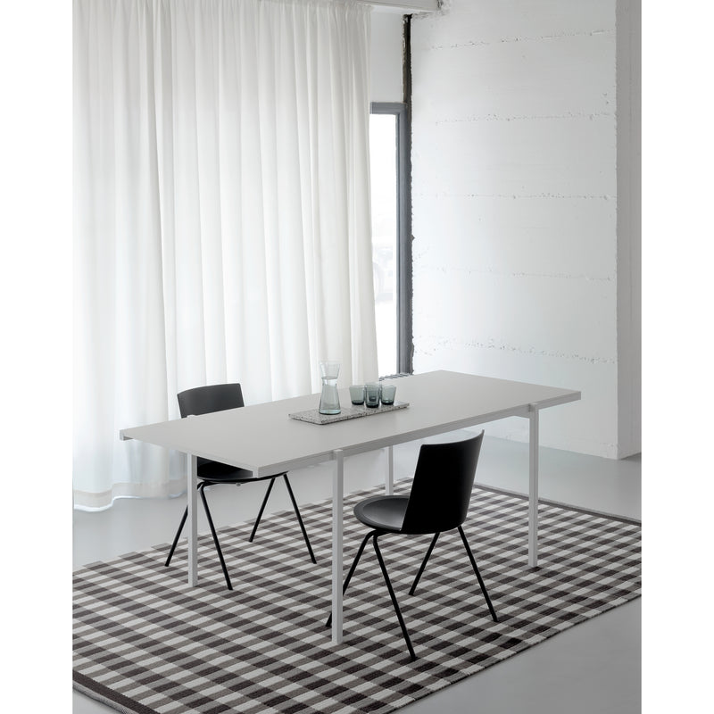 A minimalist dining area showcases a DL5 Neo Table by Loehr with steel tubes, flanked by two black chairs and topped with glassware. A checkered rug lies beneath near a window adorned with white curtains, reflecting the De Stijl movements clean modern aesthetic of white walls and floors.